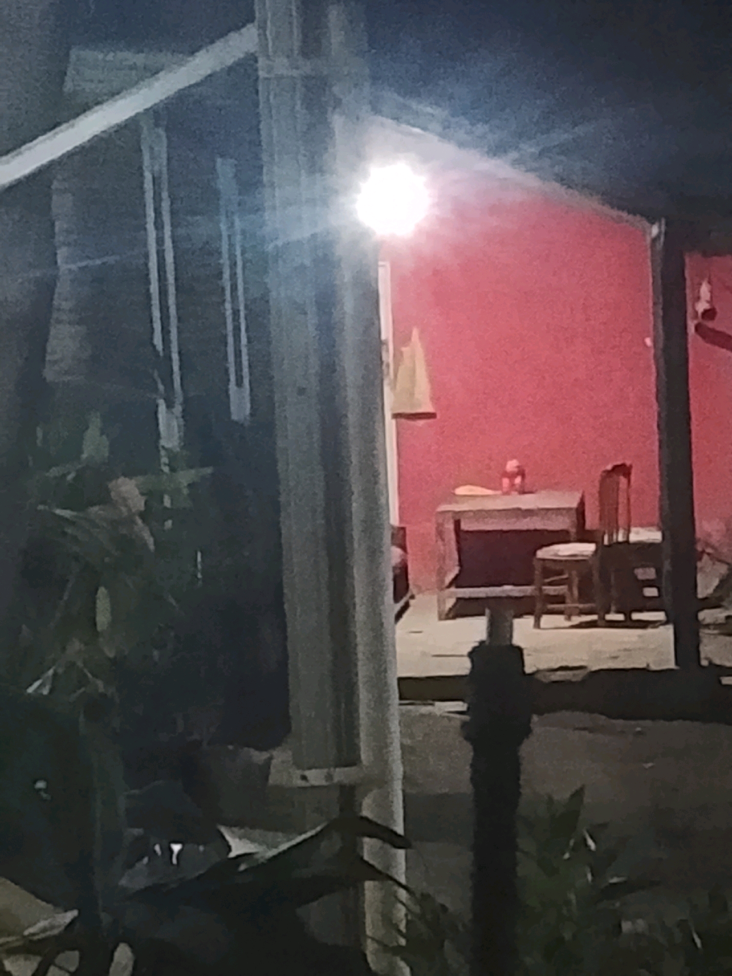The image captures an outdoor scene at night in Ujungrusi, Indonesia, likely a residential area or a small establishment. In the background, a bright light illuminates a wall painted a vibrant red. In front of this wall, a dark-colored table stands, with two small, red objects on its surface that appear to be fruits or decorations. To the right of the table, a wooden chair with a dark frame and a lighter seat is positioned. A dark, silhouetted figure is partially visible in the foreground to the left, partially obscured by lush green foliage. Another dark, vertical structure, possibly a post or pillar, is located in the middle of the frame, and a smaller, dark object rests on the ground in front of it. The overall impression is one of a dimly lit, tropical setting.