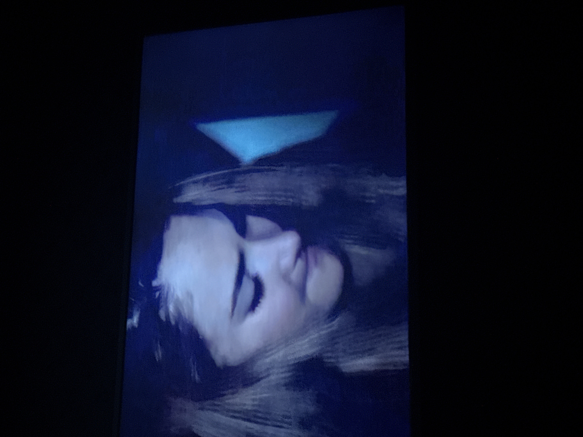 The image appears to be a screen displaying a depiction of a woman with light skin and long hair, lying down with her eyes closed. Above her head is a light-blue triangular shape. The surrounding environment is dark. There is no indication of this being in Mytishchi, Russia.