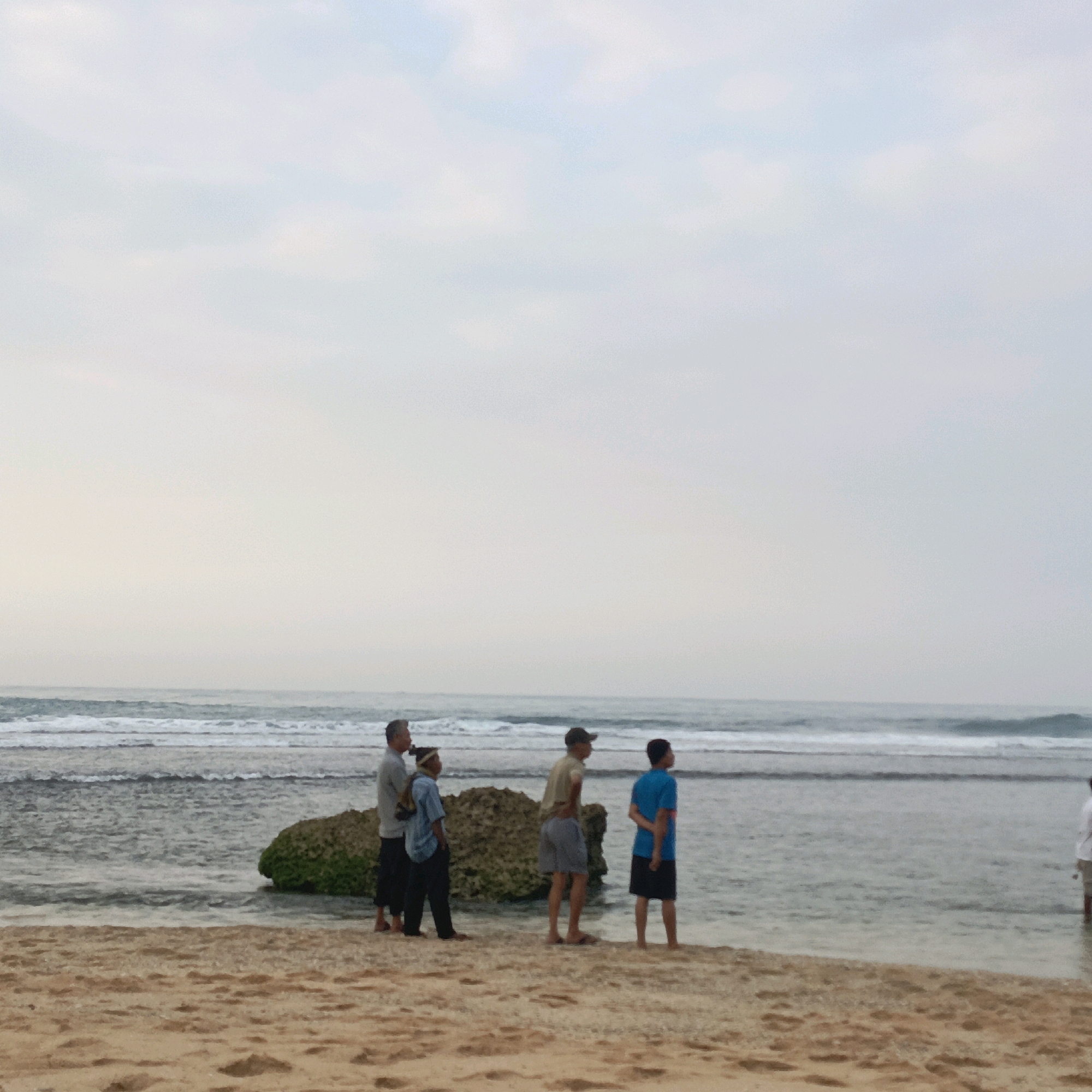 A group of five men stand on a sandy beach in Tepus, Indonesia, facing the ocean. The water is calm with gentle waves. A large, moss-covered rock sits in the shallow water to the left of the group. The sky is overcast. The man on the far left wears a grey long-sleeved shirt and dark pants. To his right, a man in a blue shirt and dark pants looks towards the ocean. The next man is in a tan short-sleeved shirt and grey shorts, with a cap on his head. The fourth man wears a blue short-sleeved shirt and black shorts. The man on the far right is partially visible and appears to be wearing a white shirt.