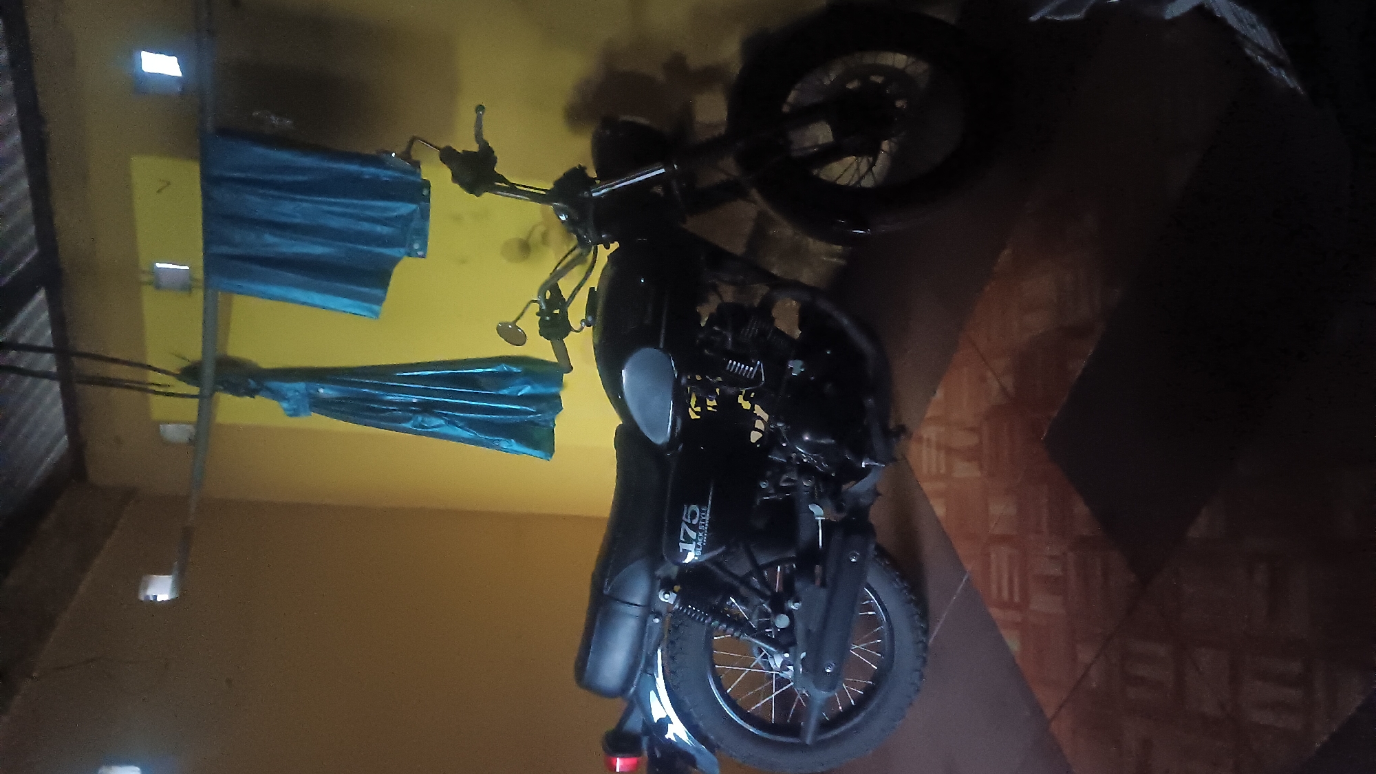 In a room with yellow walls and a corrugated metal ceiling, a black motorcycle, identified as a 175 Black Style, is parked. The motorcycle is angled to the right, with its front wheel facing away from the viewer. Two pieces of blue fabric are hanging from a bar above the motorcycle. The floor is tiled with a patterned design. The scene is lit, with shadows cast by the motorcycle and the hanging fabric. The overall impression is of a storage or display area for the motorcycle.