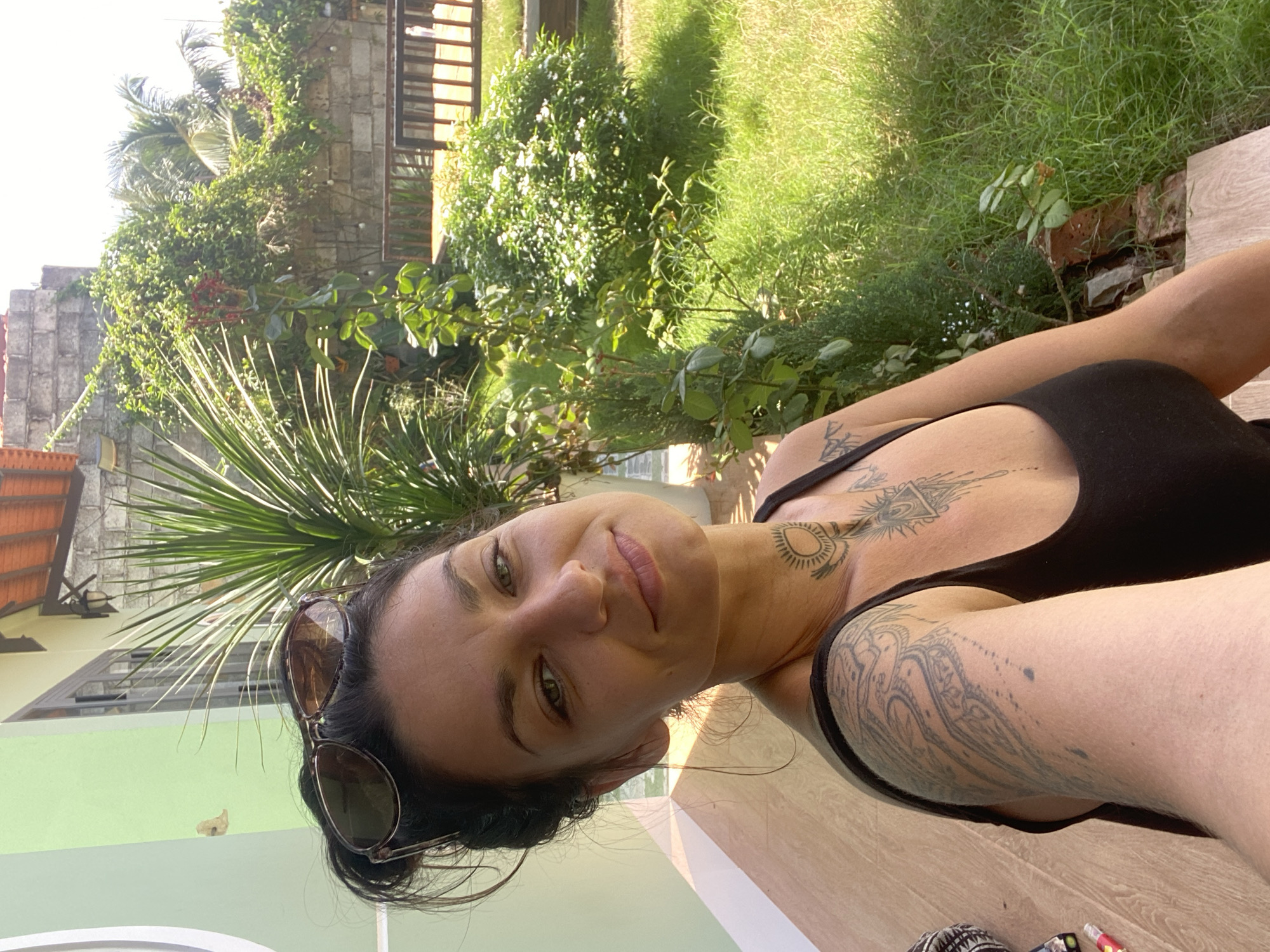 A woman with dark hair pulled up and sunglasses perched on her head sits on a wooden bench outdoors in what appears to be a residential area in Mũi Né, Vietnam. She is wearing a black tank top and has numerous tattoos on her arm and neck. The background consists of a green building, lush vegetation including palm trees and flowering bushes, and a stone wall. A wooden bench is visible in the distance. The lighting suggests it is daytime, with sunlight casting shadows.