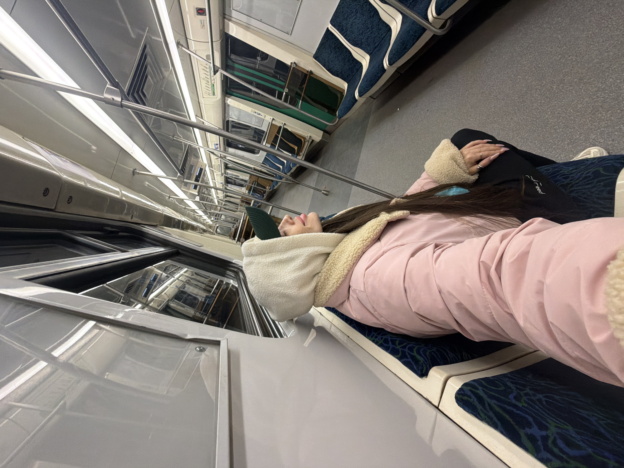 A young woman lies horizontally across two unoccupied seats in a Saint Petersburg metro car. She is wearing a pink puffer jacket with a cream-colored faux fur trim and a green baseball cap over a white hooded sweatshirt. Her black leggings, with the word "Breed" printed in white, extend to her white sneakers. The train car is brightly lit with fluorescent lighting. The interior features gray metal surfaces and navy blue fabric seats with an abstract pattern.