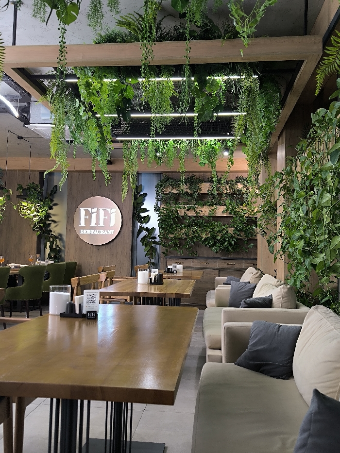 The interior of a restaurant is adorned with lush greenery. Plants cascade from a square, light-filled structure above. A vertical garden complements the wall behind a row of beige sofas with gray pillows. Wood tables with black stands are placed near the seating area. A circular logo for "FiFi Restaurant" is visible on the wall. The space is modern and inviting.