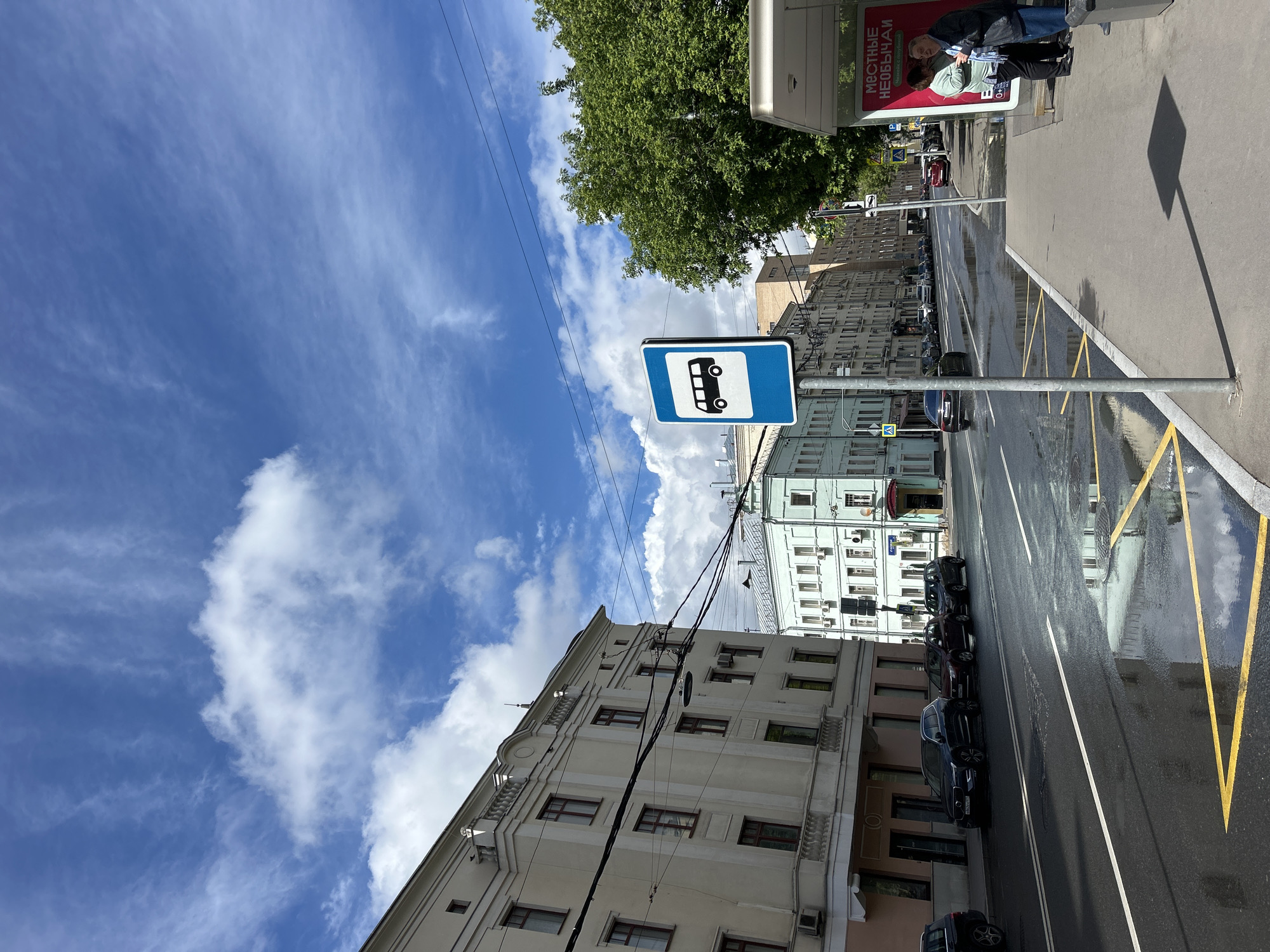 The image depicts a street scene in an urban environment. The street is wet, reflecting the surrounding buildings and sky. On the right side of the road, there is a bus stop sign, and yellow lines are painted on the curb. Several cars are parked along the left side of the street. Buildings with multiple windows and architectural details line both sides of the street. The sky is blue with scattered clouds. Power lines run above the buildings.