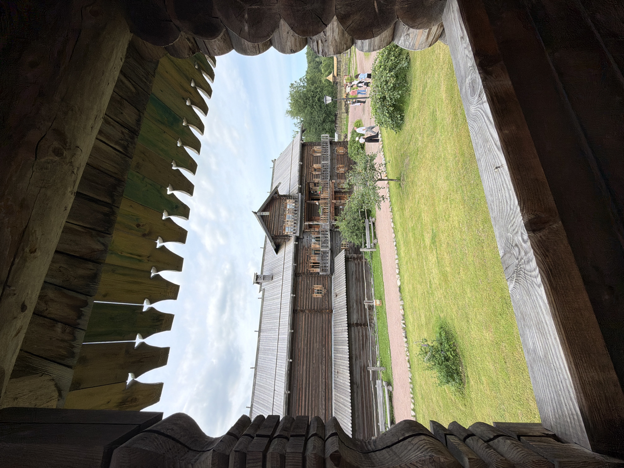 The image shows a view from a wooden porch through decorative carved eaves of a wooden building. In the foreground, the textured, aged wood of the porch structure is visible on the left and right edges, framing the scene. A weathered wooden railing runs across the bottom of the frame. Beyond the porch, a large, multi-story wooden building with a gabled roof, numerous windows, and balconies is the central subject. A pathway made of gravel and small stones leads to the building, bordered by a neatly mowed green lawn. In the middle ground, several people are walking on the gravel path in front of the wooden building. Some are gathered near a lamppost. The background consists of lush green trees and a cloudy sky. The location is identified as Nevskiy Parkleskhoz, Russia.
