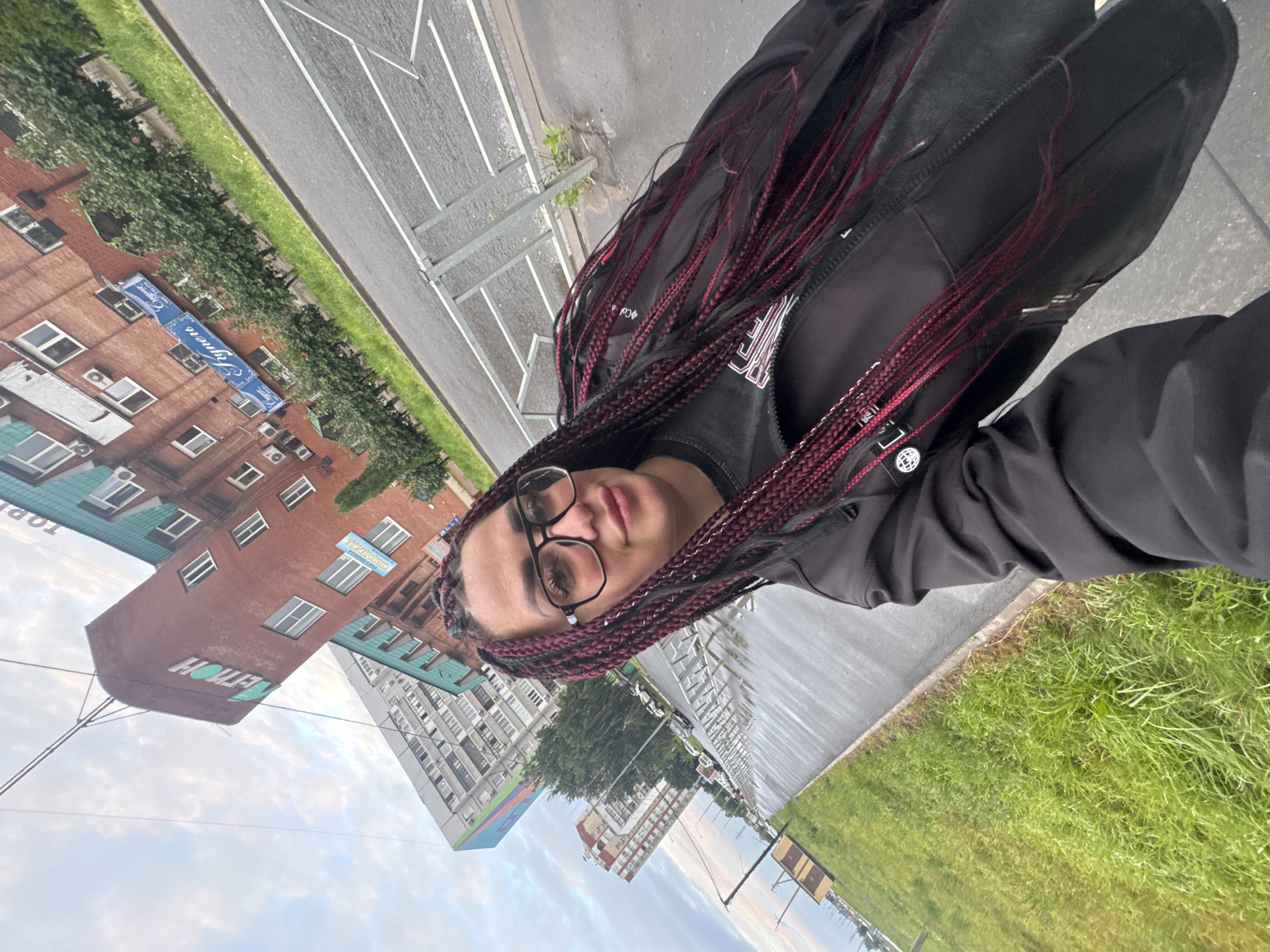 A woman with long, dark red braids and glasses is taking a selfie outdoors. She is wearing a black jacket over a dark shirt. The background shows a city street in Tolyatti, Russia, with brick buildings and a modern apartment complex. The sky is overcast, and the street appears wet, suggesting it may have rained recently. There is a metal fence along the sidewalk.