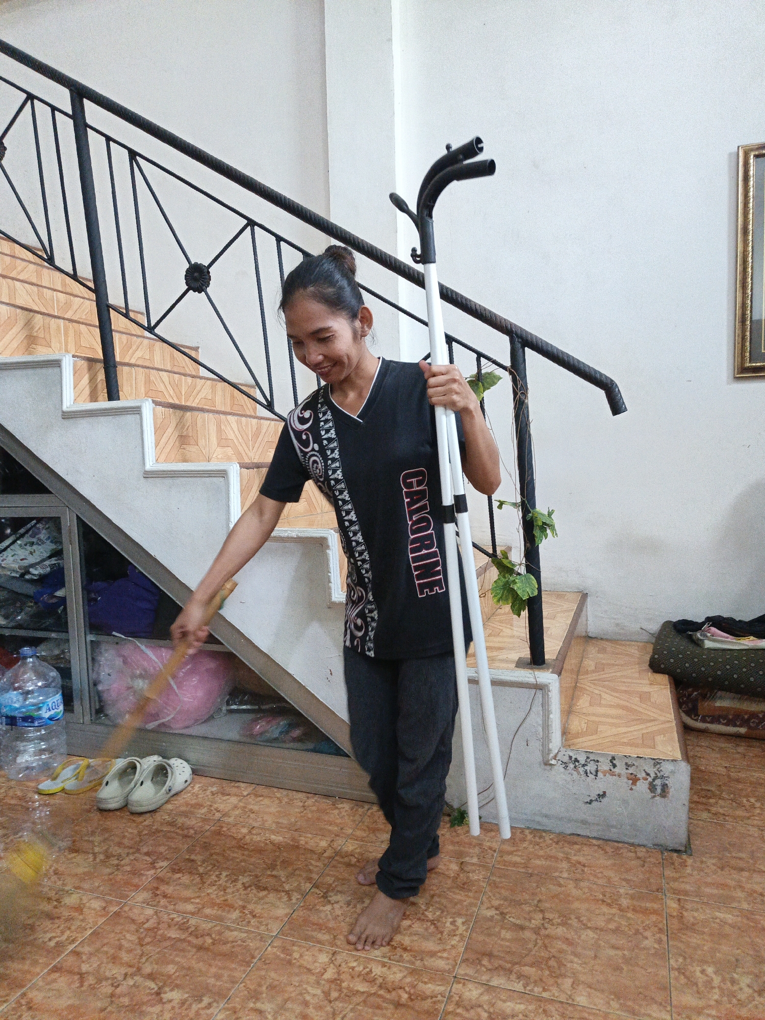 A young Indonesian woman with dark hair pulled into a bun is sweeping the tiled floor in what appears to be the entrance or common area of a residence in Karangpakel, Indonesia. She wears a black t-shirt with a decorative white and red pattern across the chest and dark pants. In her right hand, she holds a broom made of wood and straw, which she is using to sweep. She is looking down and smiling slightly as she sweeps. In her left hand, she holds a white garment rack with two curved ends, resting it against the stairs. Behind her, a set of stairs with a black metal railing ascends, with potted plants on the landing. To her left, a large bottle of Aqua water and a pair of yellow flip-flops and white clogs are visible near a glass-fronted cabinet filled with various items, including clothing and bags. The floor is tiled with orange and brown tiles, and the walls are painted white. A framed picture hangs on the wall to the right.