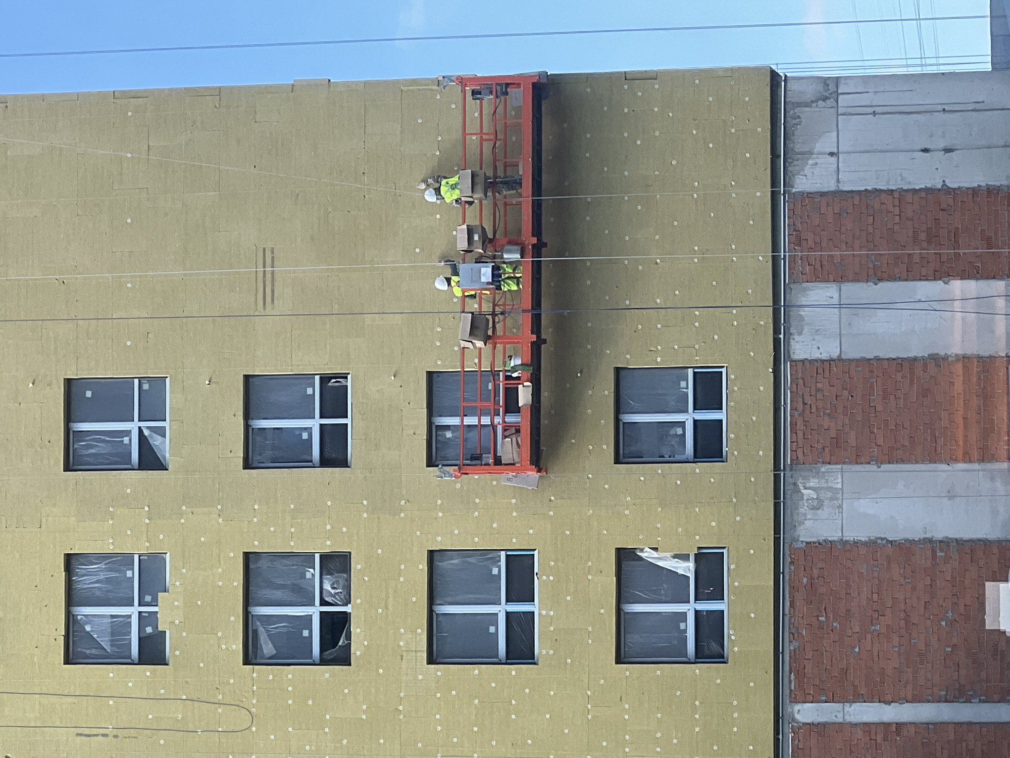 The image depicts a construction scene on the side of a multi-story building, likely located in Yekaterinburg, Russia. The building's facade is covered with yellowish insulation panels, with multiple windows visible. Two construction workers, wearing safety vests and hard hats, are positioned on a suspended platform. They are actively engaged in work, with one appearing to be placing boxes or materials, and the other holding a tool, possibly a paintbrush or applicator, and reaching towards the wall. The platform is equipped with various items, including boxes, a bucket, and what appears to be a water cooler. Below the insulated section of the building, a lower portion is constructed with red brickwork and concrete elements. The sky above is clear and blue, suggesting fair weather.