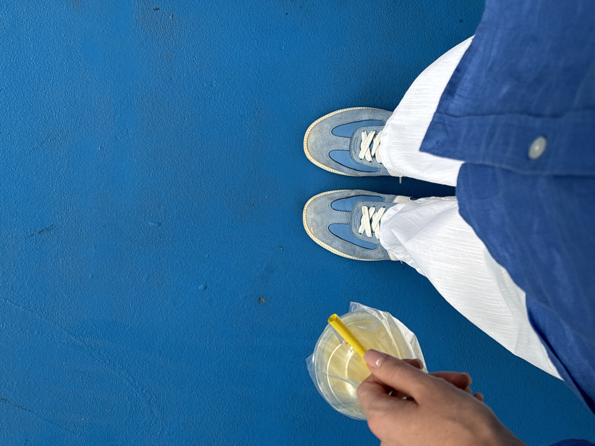 A top-down, medium shot shows a person standing on a blue surface in Tambon Sa Khu, Thailand. The person wears blue sneakers with white laces, white pants, and a blue shirt. The person holds a clear plastic cup with a yellow straw. The background is a solid blue surface.