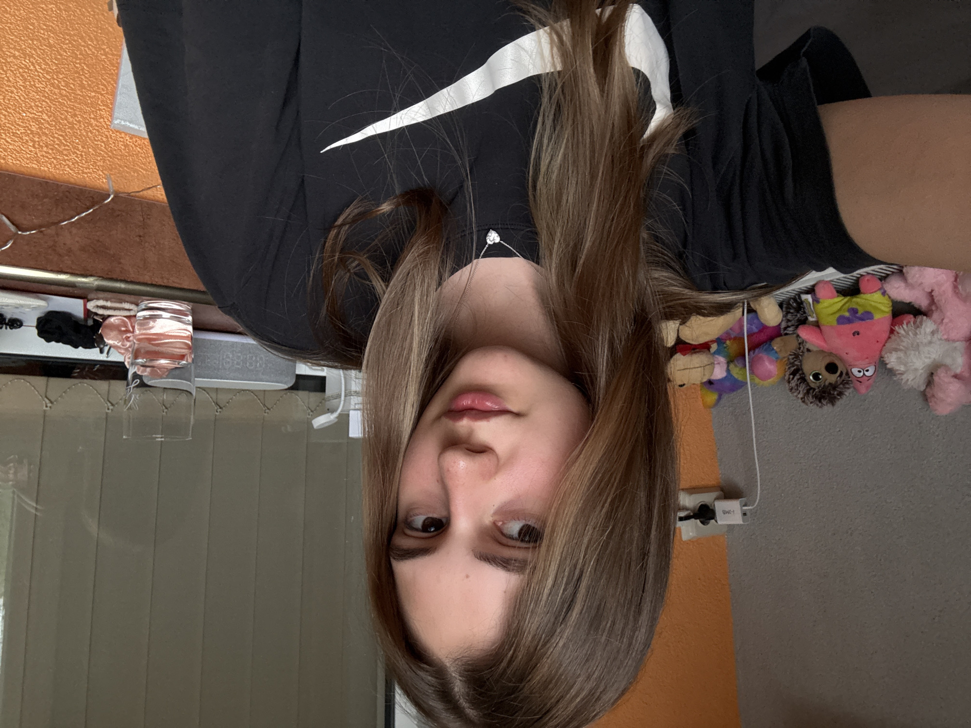 An indoor portrait features a young woman looking directly at the camera with a neutral expression. She has medium-length brown hair and is wearing a black long-sleeved shirt with a prominent white Nike swoosh logo on the chest. A delicate silver necklace with a heart-shaped pendant adorns her neck.
The background reveals a personal, lived-in setting, likely a room in a home in Panagyurishte, Bulgaria. To her left, a clear glass holding a light-colored liquid sits on a surface, next to a digital clock displaying "19:08", indicating the time is early evening. Below these items, a window with vertical blinds is partially visible. To her right, a colorful assortment of stuffed animals is arranged, including a pink pig-like toy, a green and yellow Spongebob character, a hedgehog, and a white fluffy toy. A white power adapter, labeled "SWE-1MB", is plugged into a wall outlet. The walls in the background are textured, with a vibrant orange section meeting a muted gray area.
