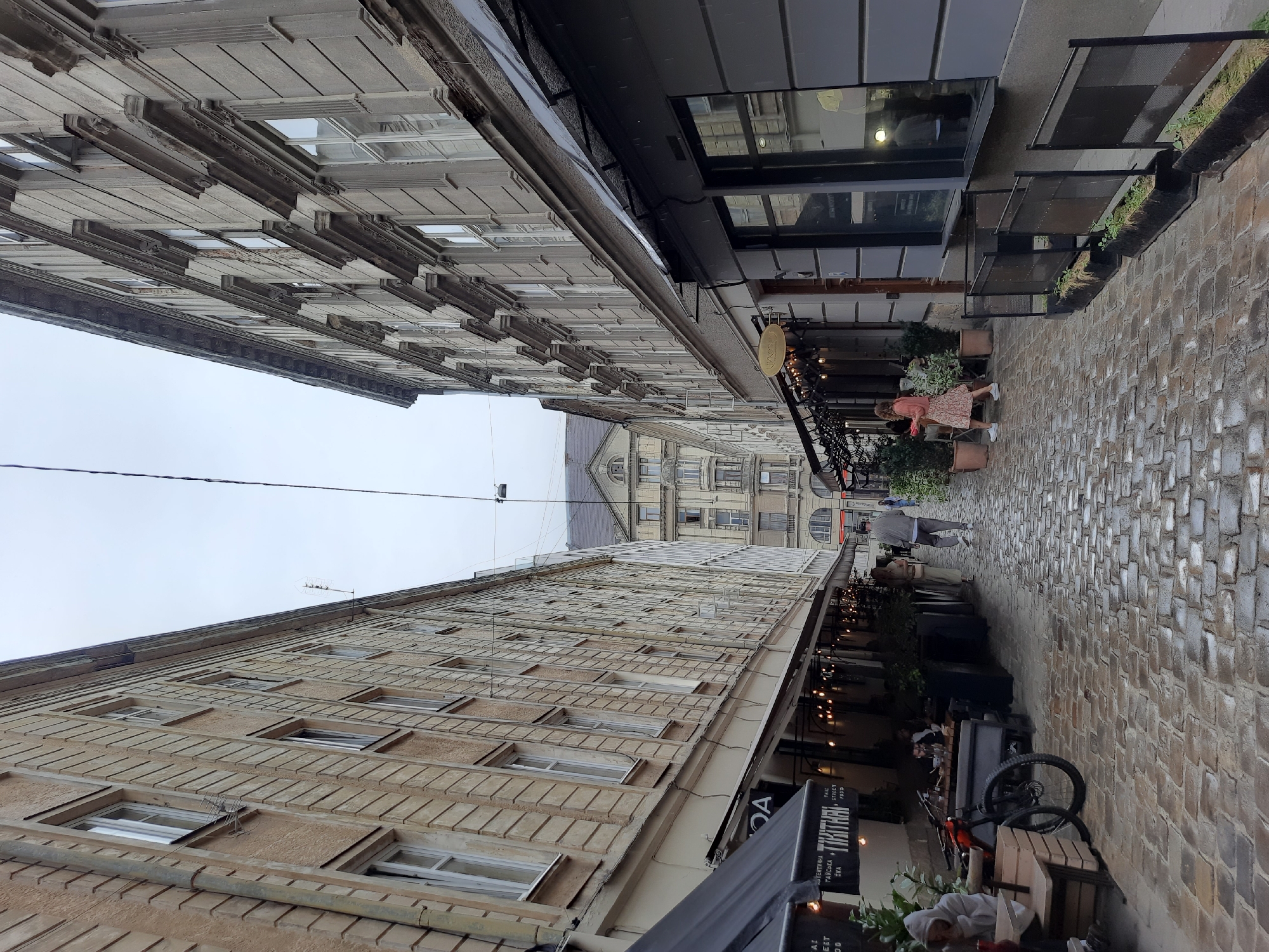 The image shows a narrow cobblestone street in Lviv, Ukraine, likely a pedestrian alleyway lined with buildings housing cafes and shops. On the left, a building with beige stucco and ornate stone detailing has awnings extending over outdoor seating areas for cafes. Lights are strung along the awnings. A bicycle is parked near a wooden crate and a seated person wearing a hooded jacket. On the right, a building with grey facade and similar architectural details also has awnings and outdoor seating. A woman in a floral dress is walking away from the camera. Further down the street, the buildings recede and several people are visible walking. The cobblestone pavement appears wet, suggesting recent rain. The sky is overcast.