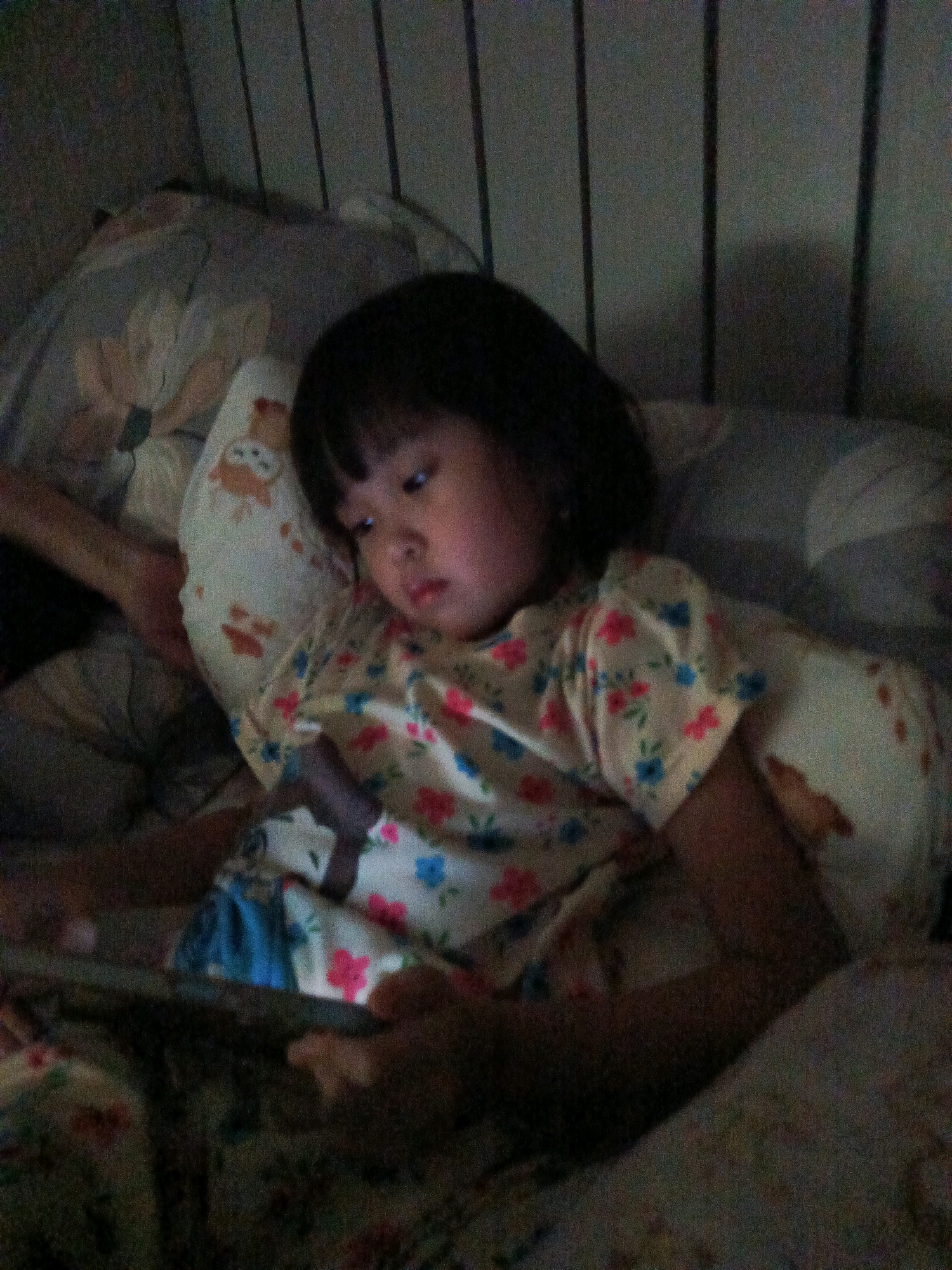 A young child is lying in bed, looking at a tablet. The child is wearing a floral-patterned pajama top and is propped up on pillows. The room is dimly lit, with vertical blinds visible in the background. The child's face is illuminated by the light from the tablet. The scene appears to be in a bedroom, possibly in a home in Curug Sangereng, Indonesia.