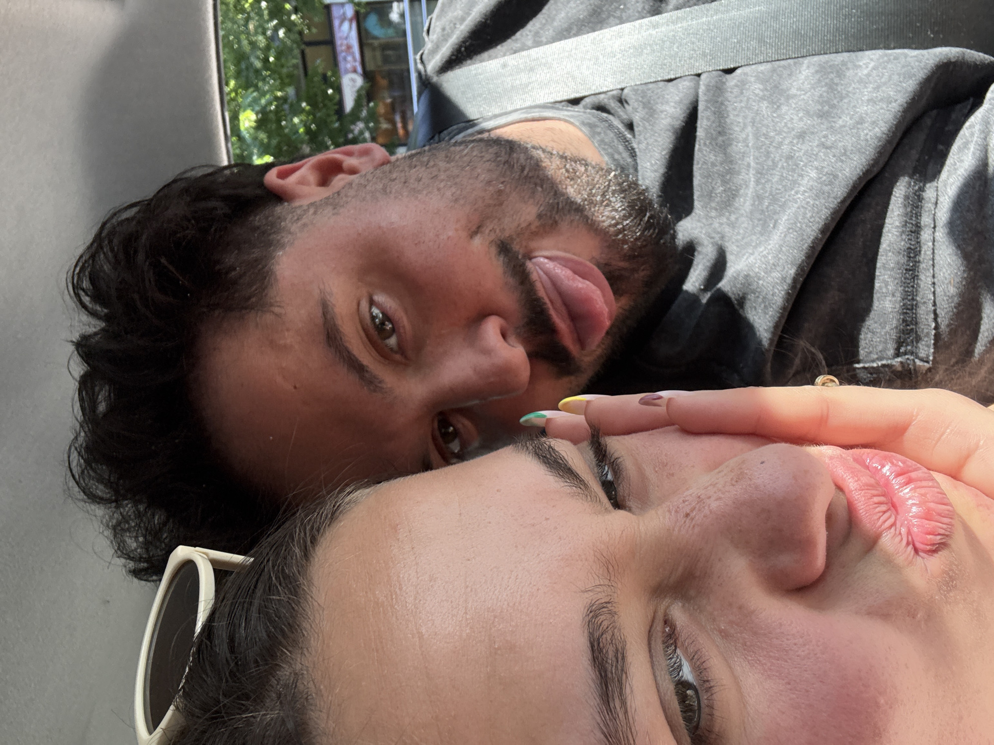 This close-up, dynamically angled image captures two individuals in a vehicle in Milan, Italy, under bright natural sunlight, indicating daytime.
In the upper left, a man with dark hair and a beard looks towards the viewer, playfully sticking out his tongue. He wears a dark grey t-shirt or sweatshirt, and a light-colored seatbelt diagonally crosses his chest. Behind him, outside the car window, are glimpses of green foliage from trees and parts of an urban building, suggesting a city environment.
In the foreground, occupying the lower right, is the upper half of a woman's face. She has dark eyebrows and brown eyes, and her lips appear textured. A hand with brightly colored nail polish (yellow, green, and pink/purple) is positioned near her face, possibly resting on her temple or cheek. A pair of white-framed sunglasses are visible resting on her head or hair. The overall mood conveyed is playful and affectionate. No discernible text is visible in the image.