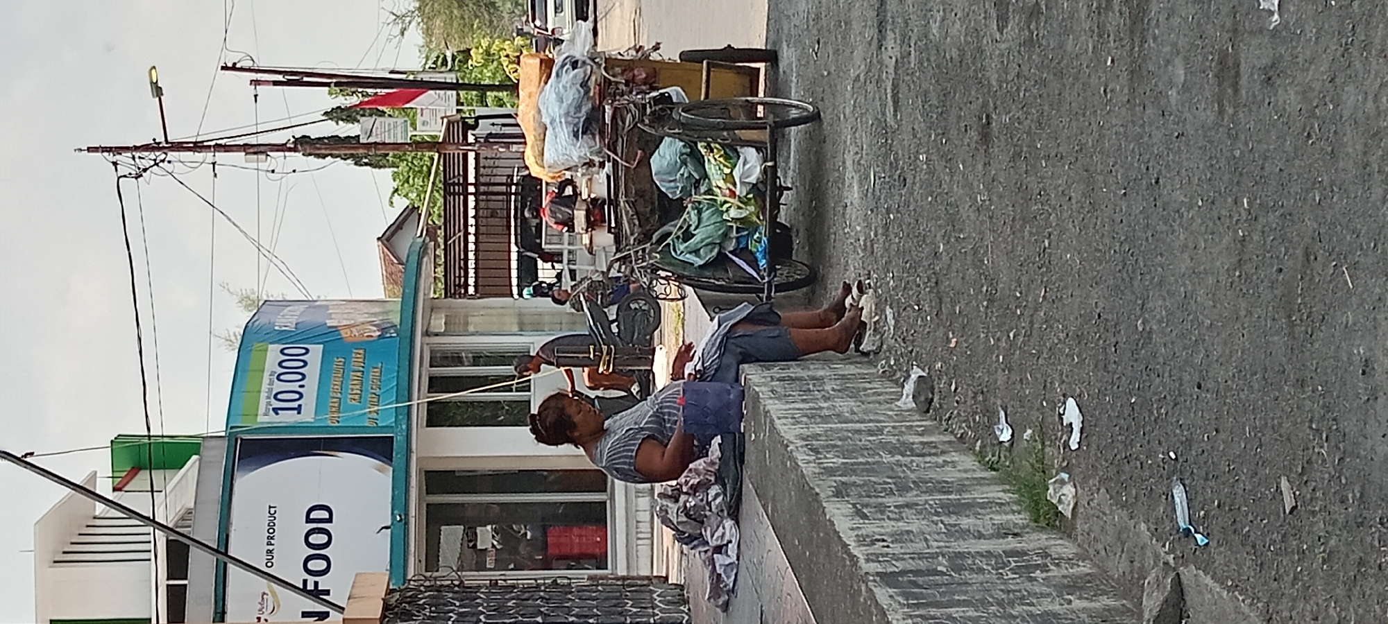 The image depicts a street scene in Tasikmalaya, Indonesia. In the foreground, a woman sits on a concrete ledge, her legs bare and resting on white material. She wears a striped shirt and dark pants, with a blue bag on her lap. To her right, a three-wheeled cart is loaded with various goods, including green fabric bags and what appear to be food items wrapped in plastic. The cart is partially obscured by the woman and her belongings. The ground is paved with asphalt and shows scattered debris, including plastic bottles and wrappers.
In the background, a building with white and green architectural features is visible, featuring a banner that reads "OUR PRODUCT" and "FOOD" along with "10.000" in large blue numerals. Several other individuals are present in the scene, some near motorcycles and a cart in front of a small kiosk. A street light is visible on a pole with numerous wires. Trees and a portion of a residential building can be seen further in the background, along with a red and white Indonesian flag. The overall impression is a moderately busy street with commercial activity and pedestrian presence.