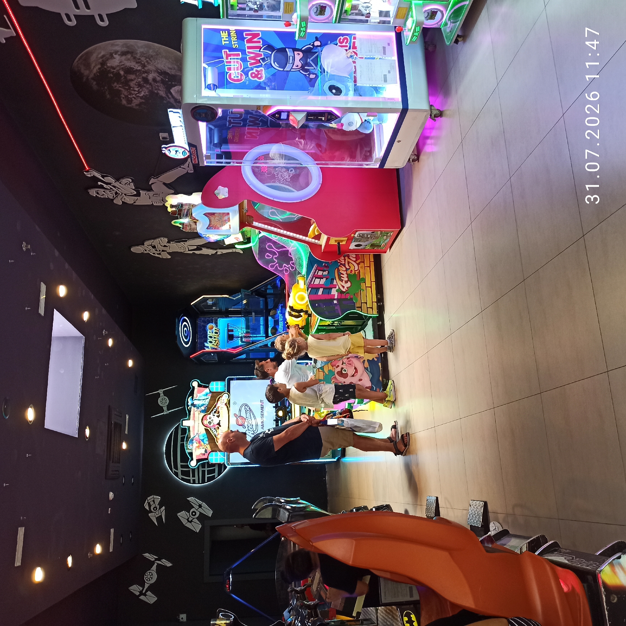 The image depicts the interior of an arcade, likely located in Modugno, Italy, as indicated by the date stamp. In the foreground, an orange, futuristic-looking arcade game seat is visible on the left. Further into the arcade, several people are present. A man in a dark t-shirt and beige shorts stands with his hands clasped, facing away from the camera, while a young boy in a light t-shirt and dark shorts stands to his right, looking towards a game. Another young child in a yellow t-shirt and shorts is also facing a game. In the background, various arcade machines are illuminated, including a "Hoop Shot" game and a crane game with a "CUT THE STRING & WIN" sign. The walls are decorated with graphics, including Star Wars-themed elements like TIE fighters and stormtroopers, as well as a large depiction of a planet. The floor is tiled, and overhead lighting illuminates the space. A timestamp "31.07.2026 11:47" is visible in the bottom right corner of the image.