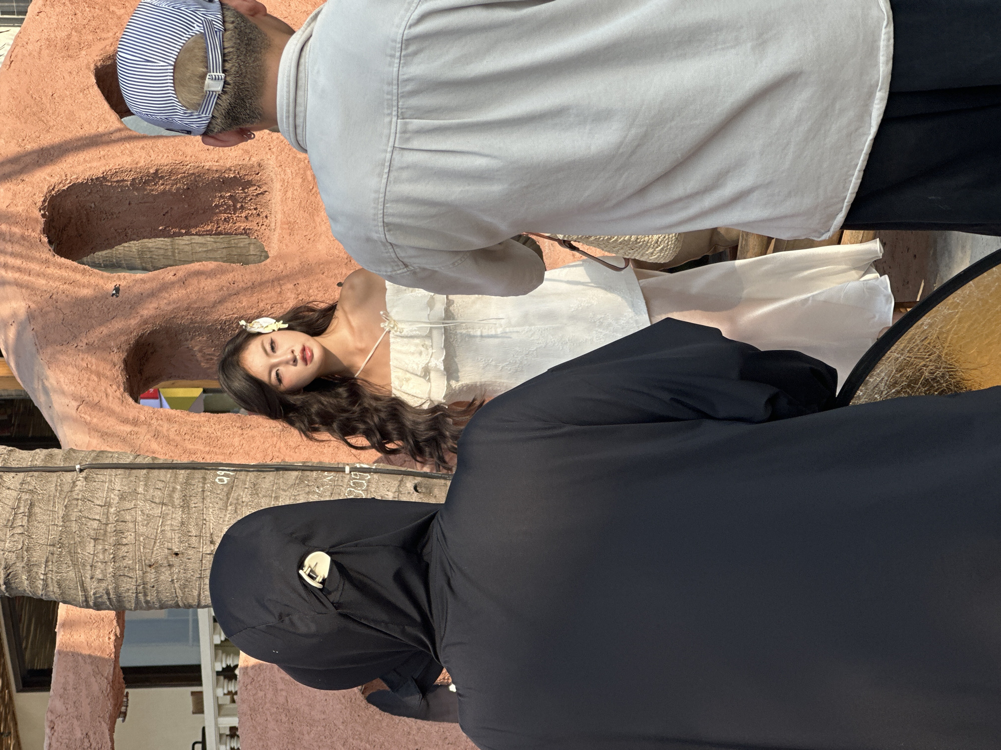 The image captures a scene in what appears to be a photography session. A woman in a white dress with a floral headpiece is positioned against a uniquely textured, reddish-brown wall with cavernous openings. Two individuals are positioned around her; one is wearing a light-colored shirt and a striped baseball cap, and the other is wearing a black hooded garment. The former appears to be the photographer, while the latter might be a lighting assistant or part of the crew, partially obscuring the view. A reflective gold disc is present, presumably for lighting purposes. The surrounding environment suggests an outdoor setting in what may be a beach resort or a similar structure, with a tree trunk visible in the scene. Based on the architectural style and environment, the location is likely in or near Đà Nẵng, Vietnam, known for its coastal resorts and unique constructions.