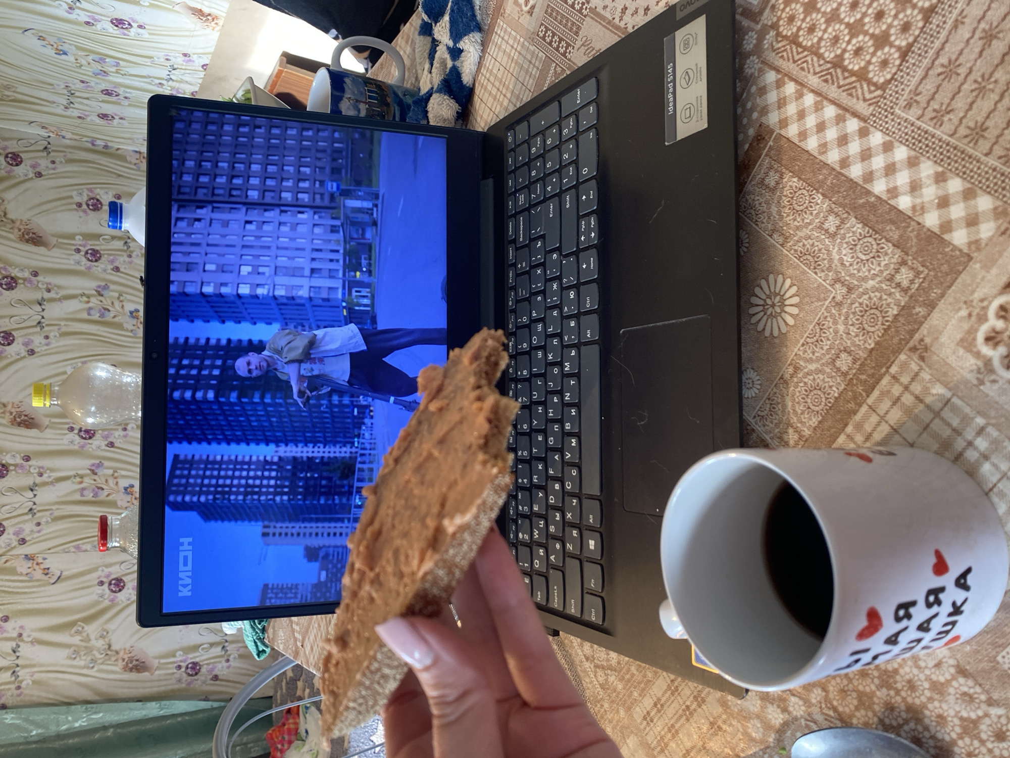 The image depicts a close-up, over-the-shoulder view of a person holding a piece of rye crispbread spread with a light brown paste, likely peanut butter, over a black Lenovo Ideapad S145 laptop. The laptop screen displays a scene with tall, modern buildings and a person standing in the street, with the logo "КИОН" visible in the top left corner of the screen. To the right of the laptop, a white ceramic mug with a design of what appears to be a European city skyline and red heart accents, and the Russian text "Моя любимая чашка" (My favorite mug) is present. A checkered blue and white cloth is also visible on the right. The setting appears to be indoors, with patterned wallpaper in the background and a patterned tablecloth on the table where the laptop and mug are placed. The overall impression is of a casual moment of watching content and snacking, likely at home. The text on the mug and the "КИОН" logo suggest a Russian context.