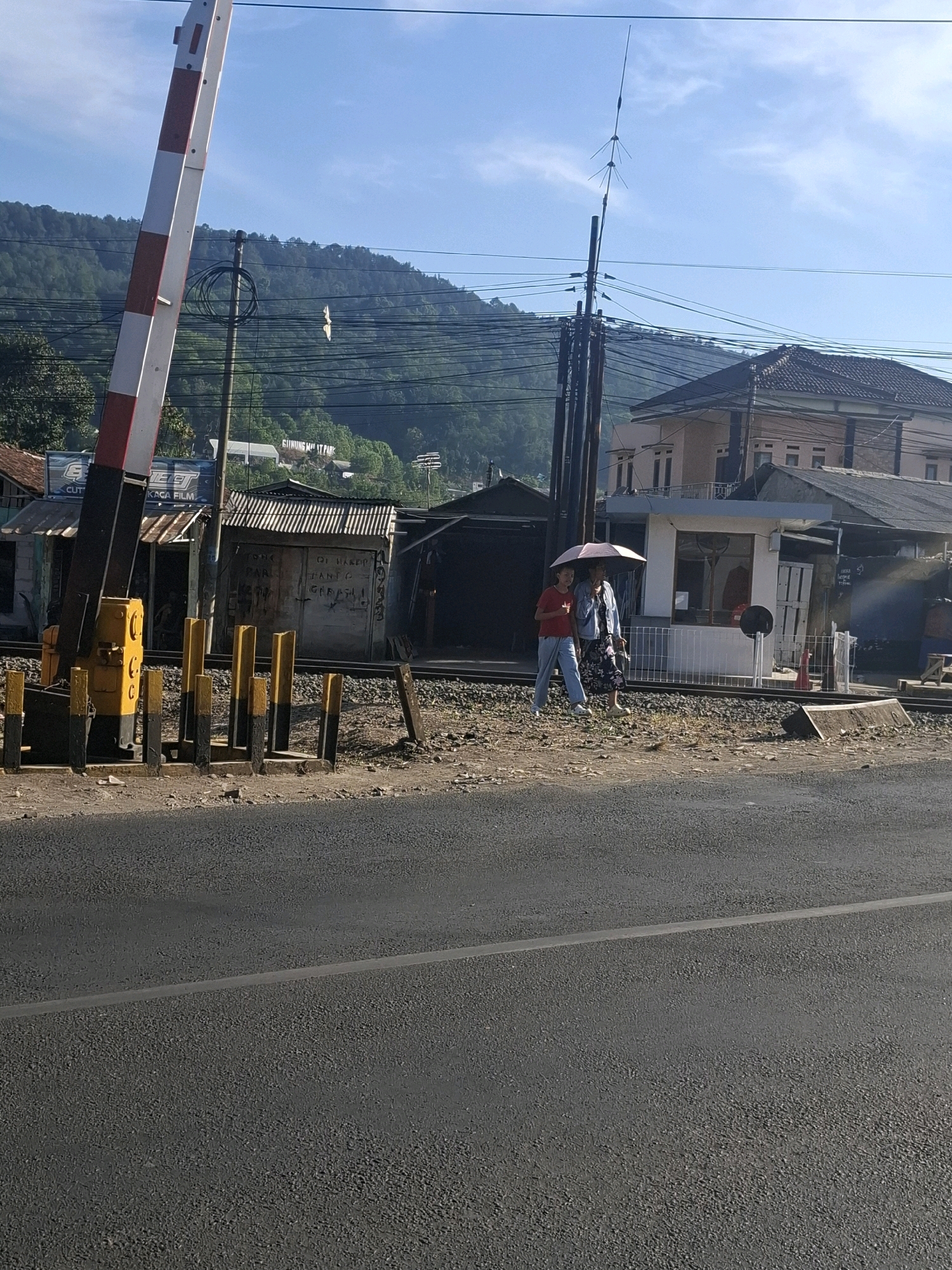 The image depicts a railway crossing with a barrier arm in the foreground, painted with red and white stripes. A paved road runs across the bottom of the frame. In the middle ground, two individuals are walking across the railway tracks, one holding a pink umbrella. Adjacent to the tracks are several small buildings and structures. A light-colored, multi-story building with a dark tiled roof is visible behind the crossing. The background features a lush, green, tree-covered hillside under a clear blue sky with scattered clouds. Utility poles and wires are strung across the scene. The location is identified as Lembursawah, Indonesia.