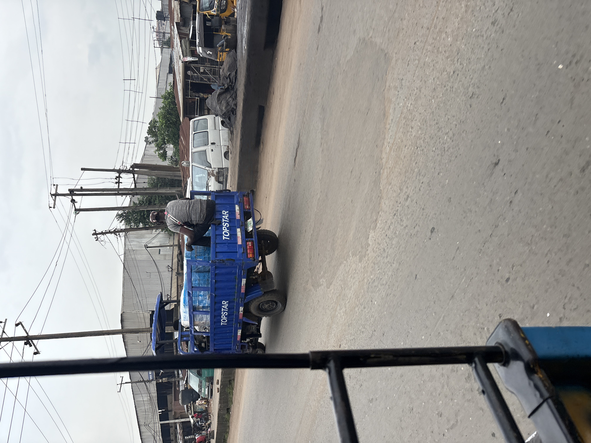 A man sits on the back of a blue truck in Onitsha, Nigeria, with his back to the camera. The truck is loaded with cases of bottled water, some of which are wrapped in plastic. The truck is on a paved road, with a white van and several other vehicles in the background. Buildings with corrugated metal roofs are visible on the left side of the road, and overhead power lines crisscross the sky. The sky is overcast. The truck is branded with the word "TOPSTAR" on its side.