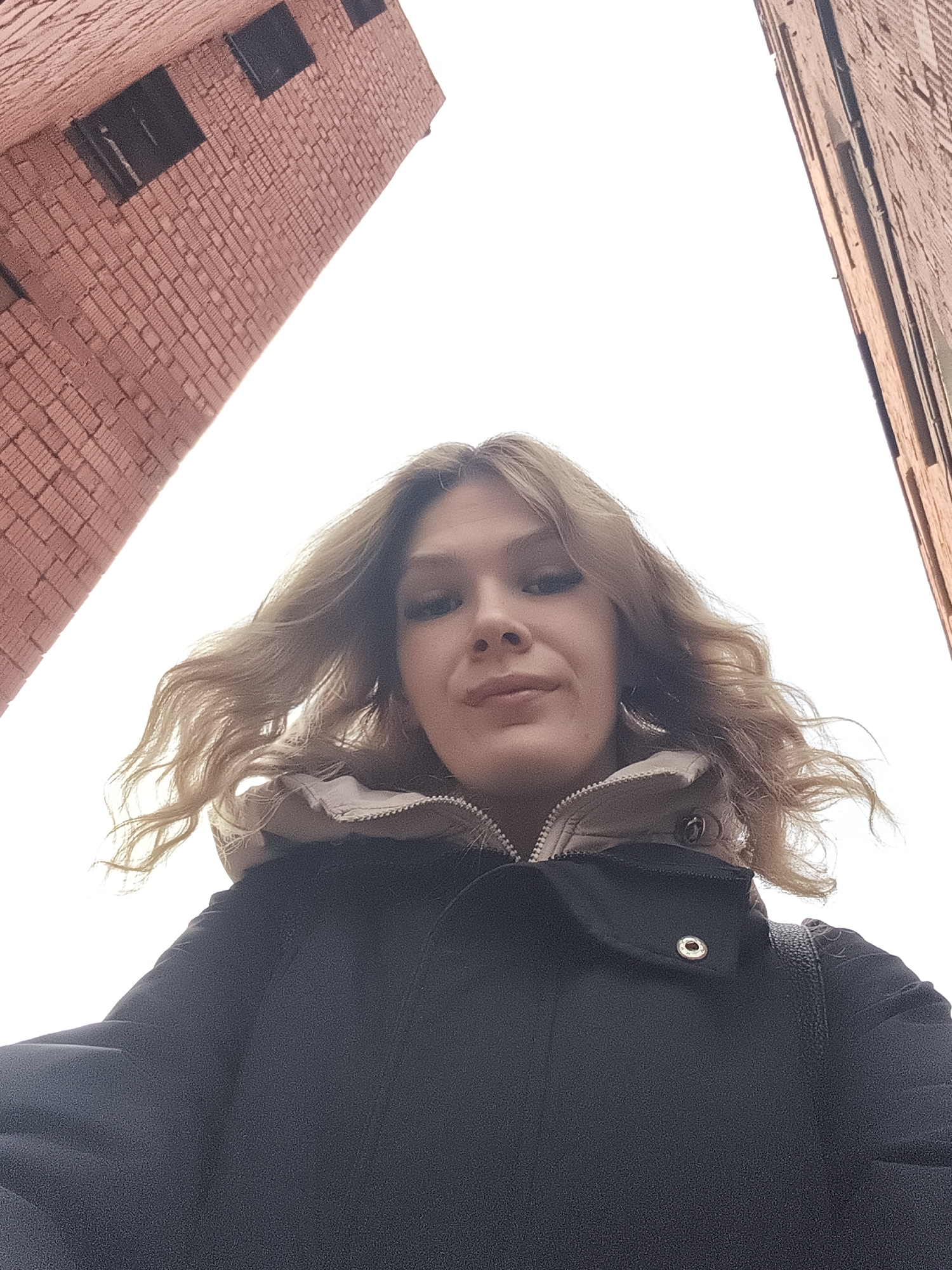 The image presents a low-angle shot of a woman outdoors. She has blonde hair and is wearing a black coat with a beige lining visible at the collar. Two tall brick buildings frame the upper part of the photo, converging towards the bright, overcast sky. The buildings appear to be located in the city of Izhevsk, Russia.