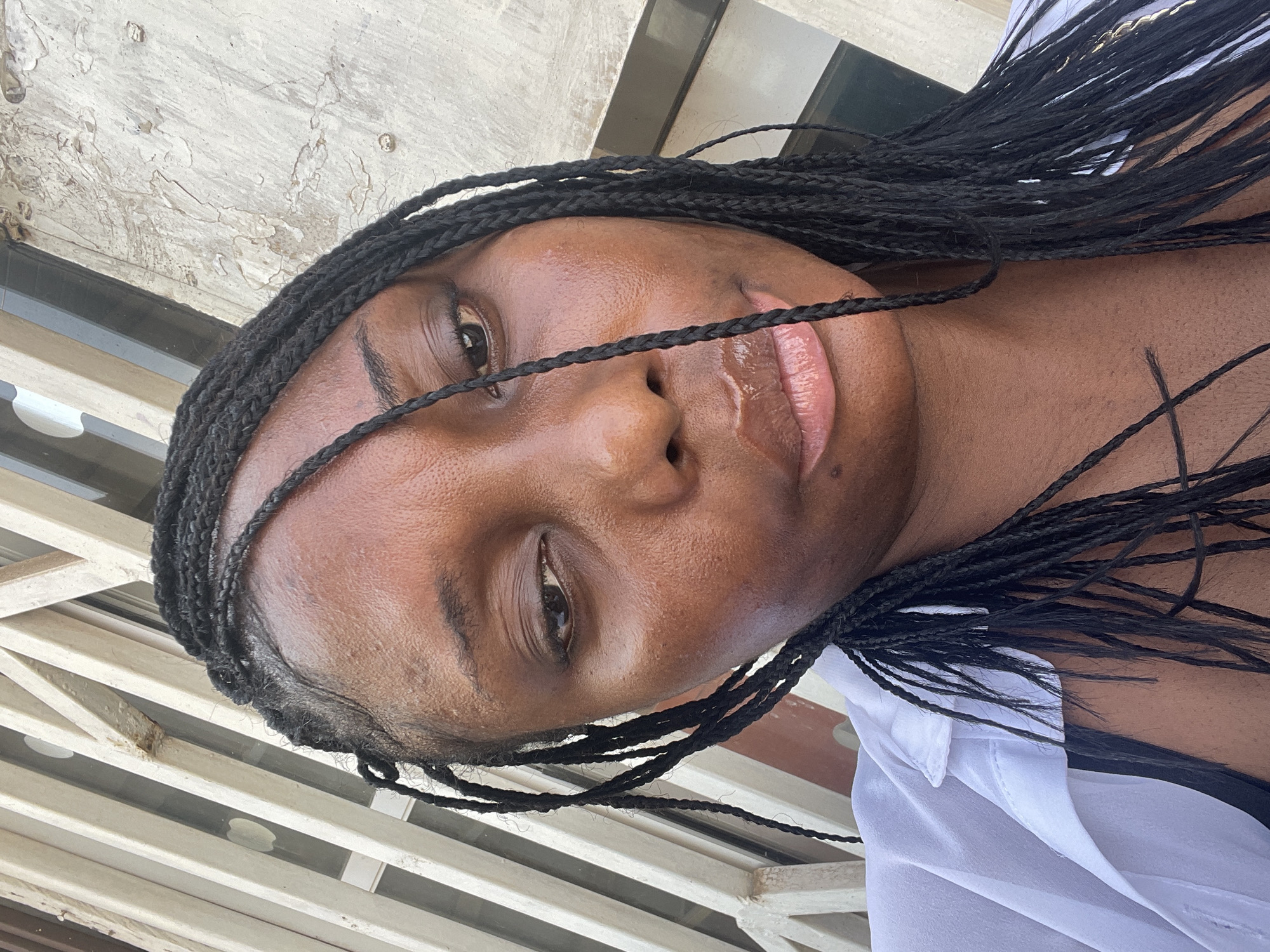 The image shows a close-up selfie of a Black woman with braided hair. She is wearing a white collared shirt over a black undergarment. Her face is turned slightly to the side, and her eyes are partially closed. The background consists of a textured, light-colored wall and vertical bars. The image does not contain information that would indicate the location is Abuja, Nigeria.