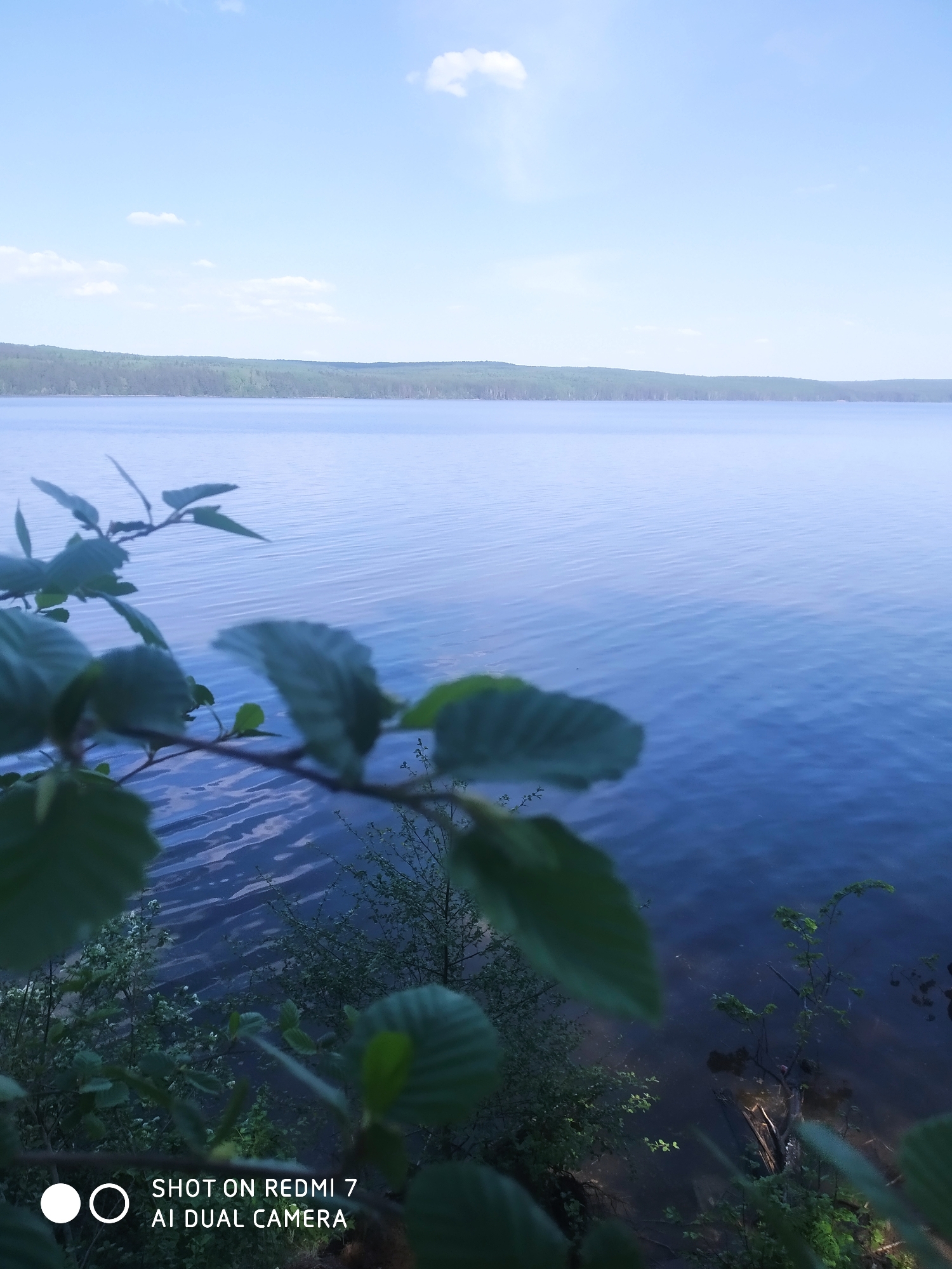 The image shows a lake in Chaikovsky, Russia. The water is calm, reflecting the sky. In the distance, there is a treeline. In the foreground, there are out-of-focus tree branches and leaves. The image was taken with a Redmi 7 AI dual camera.