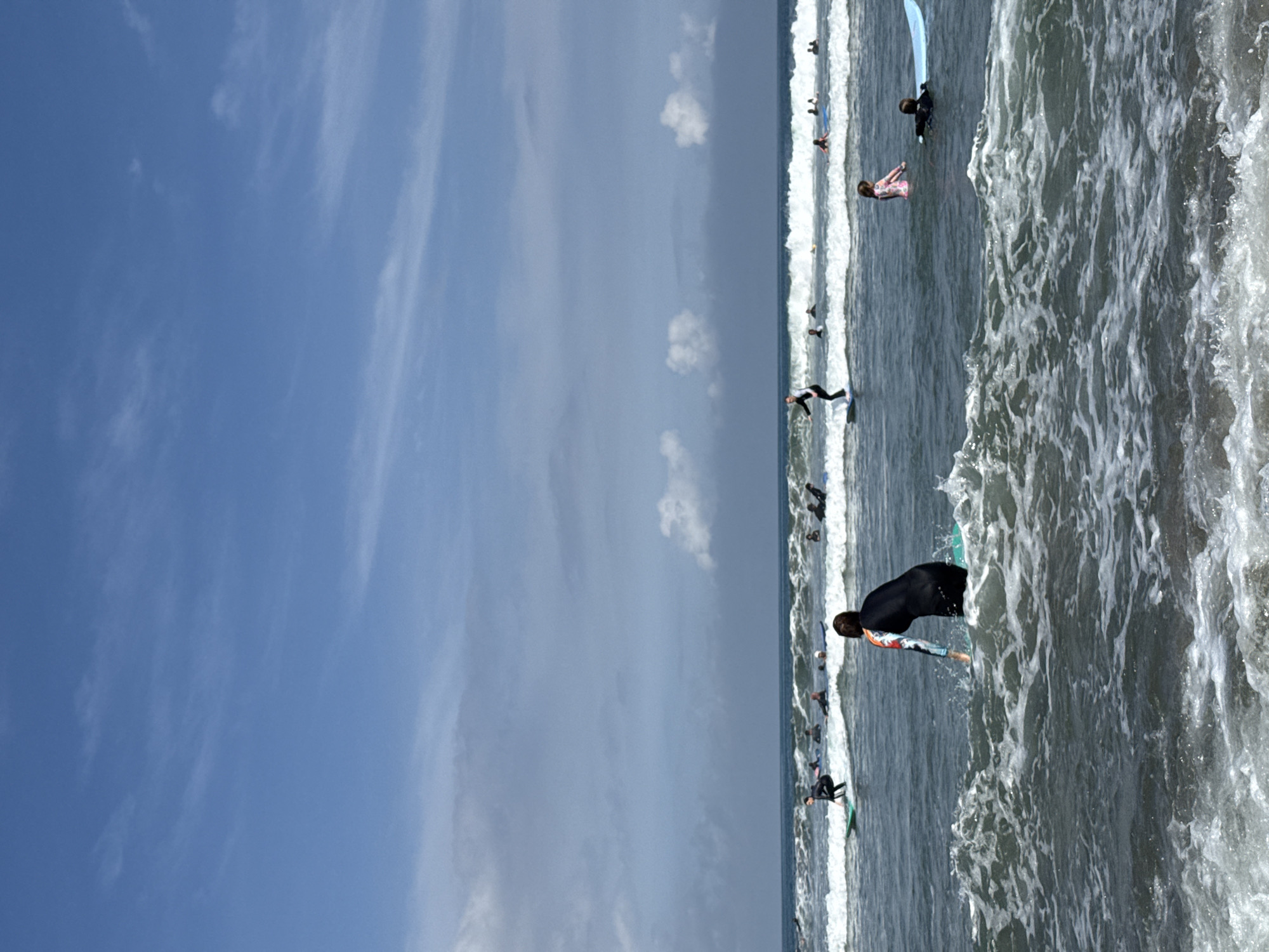 The image shows a beach scene at Kuta, Indonesia, with numerous surfers enjoying the waves. The water is turbulent with white foamy surf. Several individuals are actively surfing, while others are seen further out, waiting for waves. A clear blue sky with scattered clouds occupies the left portion of the frame. The image captures a typical day at this popular surfing destination.
