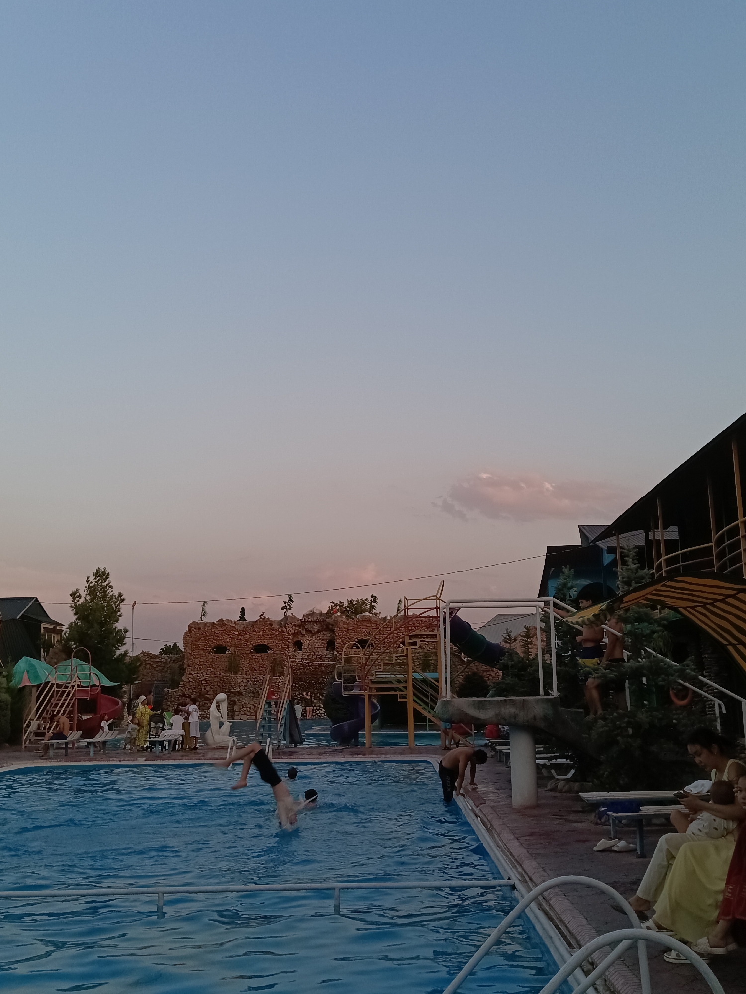 The image depicts an outdoor swimming pool area in Shymkent, Kazakhstan, likely an amusement park or water park, during what appears to be late afternoon or early evening, judging by the soft lighting and dusky sky. In the foreground, a person is captured mid-air, performing a somersault into the bright blue water of the swimming pool. The pool is large and rectangular, with rippling blue water reflecting the sky. Several figures are visible around the pool, some swimming and others lounging on the poolside.
To the right of the pool, a covered seating area with a striped awning extends outwards. People are seated in this area, including a woman holding a baby and another child. Further back, a man is seen standing at the edge of the pool. In the background, there are various structures associated with a water park, including colorful slides, climbing frames, and a decorative stone wall resembling a castle. Trees and other foliage are interspersed throughout the area. Several buildings with dark roofs are also visible, suggesting that this is a developed recreational facility. The overall atmosphere appears relaxed and familial, with people enjoying leisure time outdoors.