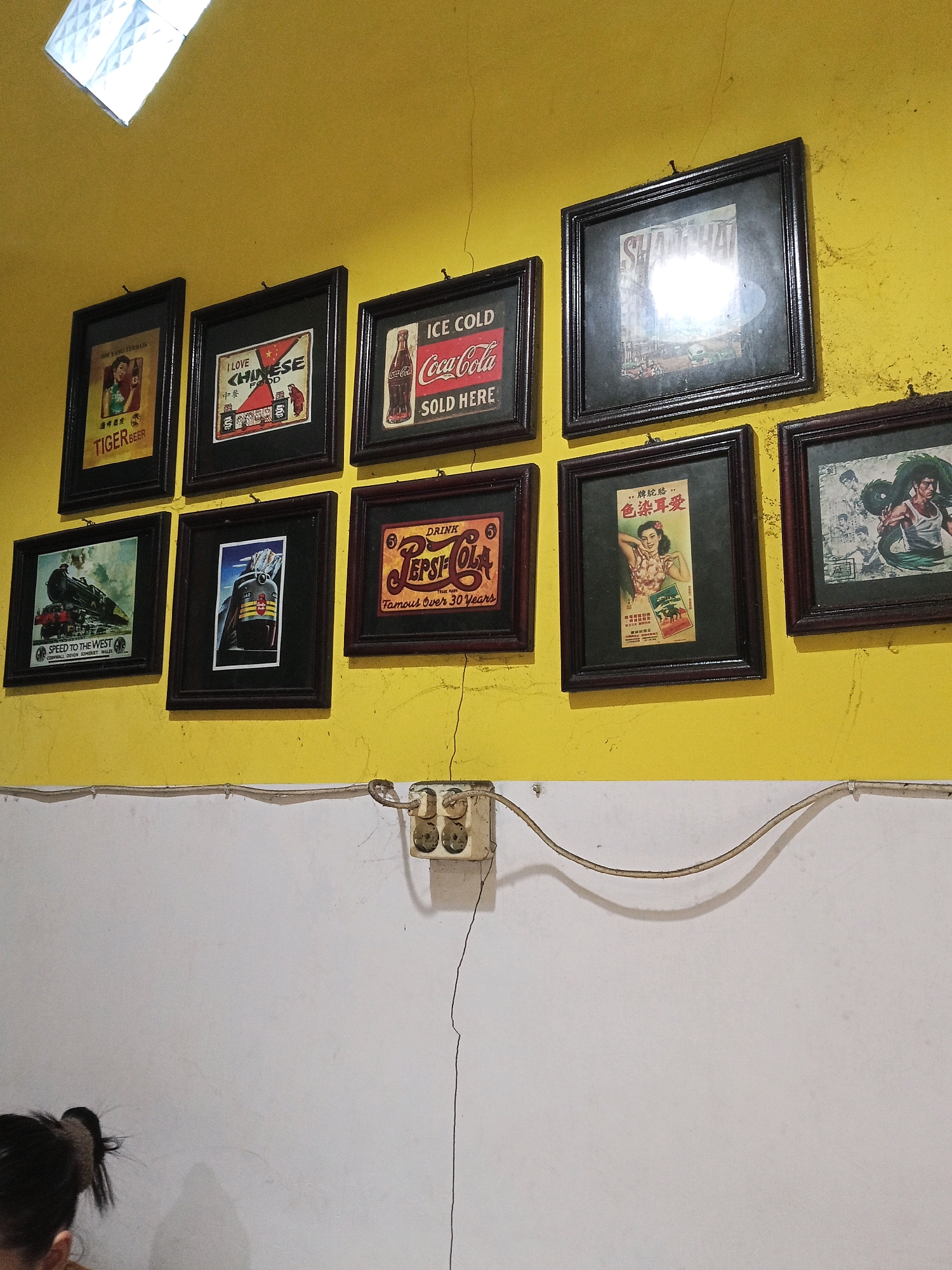 The image shows a collection of framed advertisements and artwork displayed on a yellow wall. The items are arranged in two rows. The top row contains four frames, and the bottom row contains four frames. The advertisements appear to be vintage, featuring brands like Tiger Beer, Coca-Cola, and Pepsi-Cola, as well as a vintage image of Shanghai. There are also frames with images of trains and a picture of a woman in traditional attire. Below the wall, there is a white partition, and a portion of a person's head is visible in the lower left corner. An electrical outlet with a cord is mounted on the white partition. The setting is likely a commercial establishment, possibly a cafe or restaurant, in the city of Blitar, Indonesia, given the context provided. The lighting suggests an indoor environment, possibly illuminated by overhead lights or natural light from a window, as indicated by a brightly lit square in the upper left corner of the image.