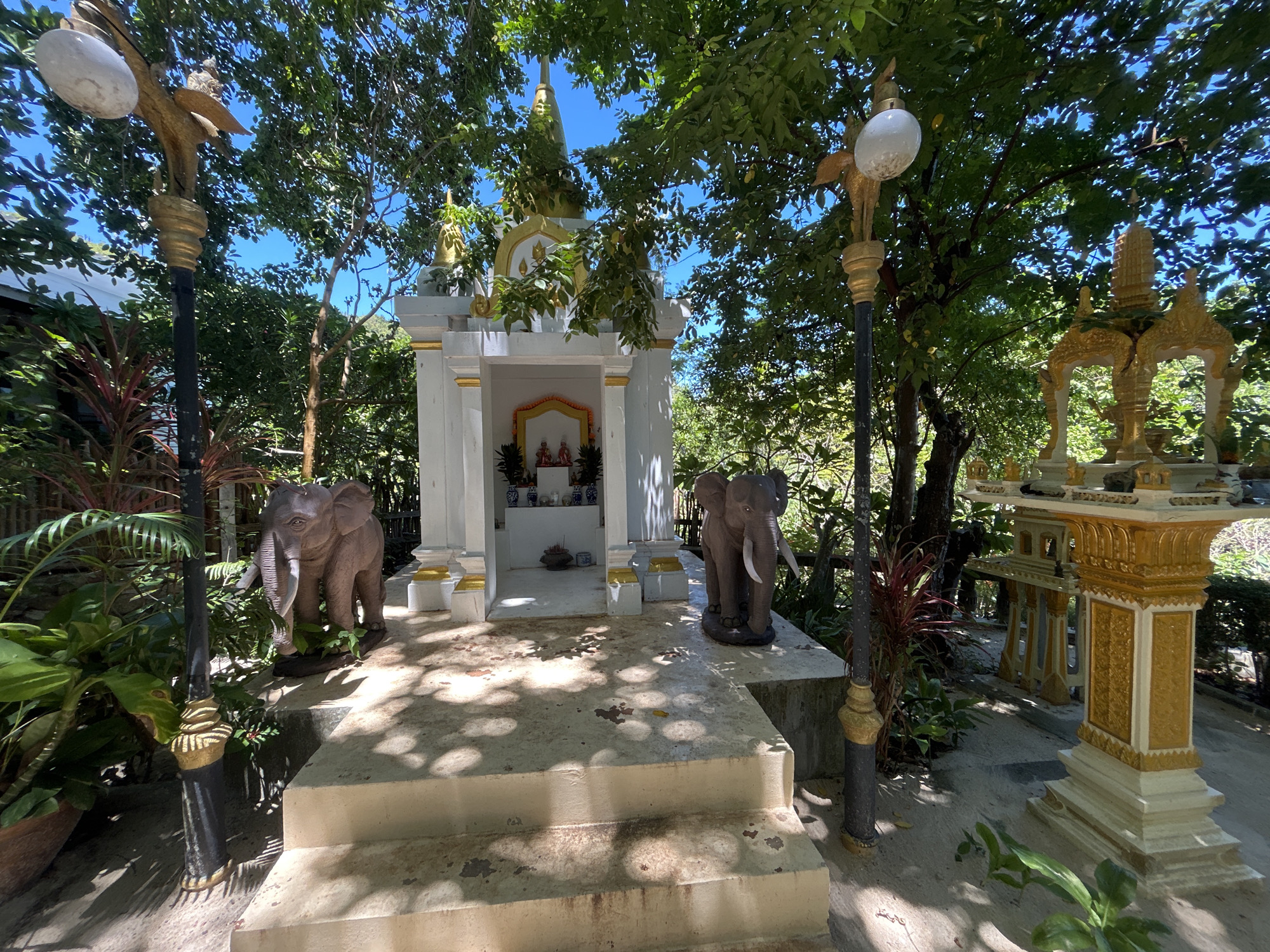 This is a wide-angle, eye-level shot of an outdoor shrine in Koh Phangan, Thailand. The shrine is white with gold accents and features a small altar inside with religious figures and offerings. Two elephant statues flank the shrine, one on each side. The area is surrounded by lush green trees and foliage, with dappled sunlight filtering through the leaves, creating a warm, serene atmosphere. Two ornate lampposts, also with gold accents, stand near the shrine, adding to the decorative elements. The ground is paved with concrete and has a stepped approach to the shrine. The overall scene suggests a place of worship or spiritual significance.