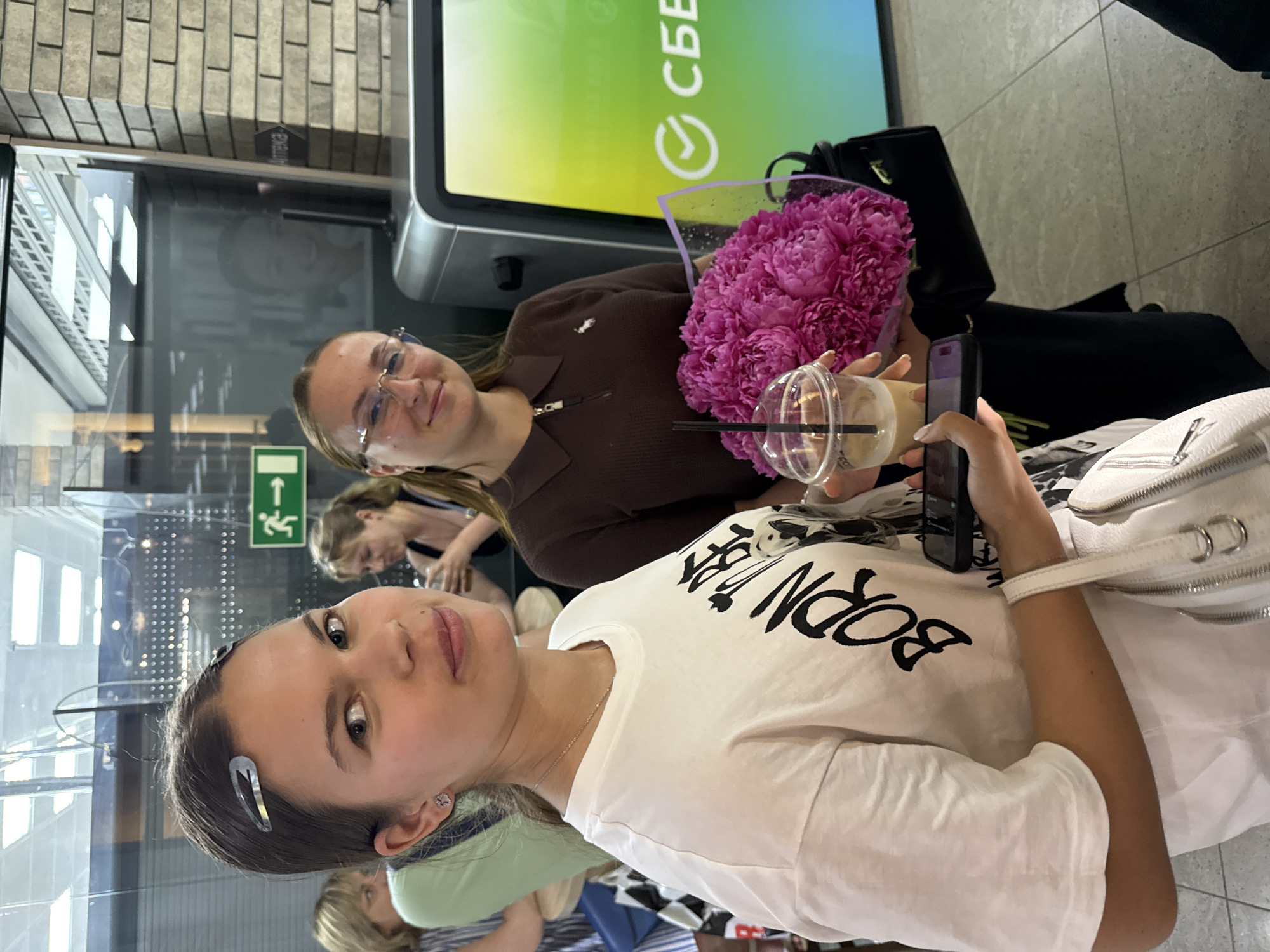 Two young women are standing in an indoor space, possibly a transit hub or a public building. The woman in the foreground on the left is wearing a white t-shirt with black lettering that reads "BORN TO BE" and has a black graphic of a face. She has a light-colored drink in a plastic cup with a straw and is holding a smartphone. She has two silver hair clips in her hair and is wearing small stud earrings. The woman on the right is wearing a brown collared shirt and glasses. She is holding a bouquet of vibrant pink peonies and a black handbag. Behind them, a large screen displays a green and white logo, possibly of a bank or company. A sign with a green directional arrow pointing to the right is visible on a glass partition in the background, indicating an exit or pathway. Another sign with Russian text and an arrow is partially visible on a brick wall. The floor is tiled. Other people are visible in the background, appearing to be seated or standing. The lighting suggests an indoor environment with overhead lights.