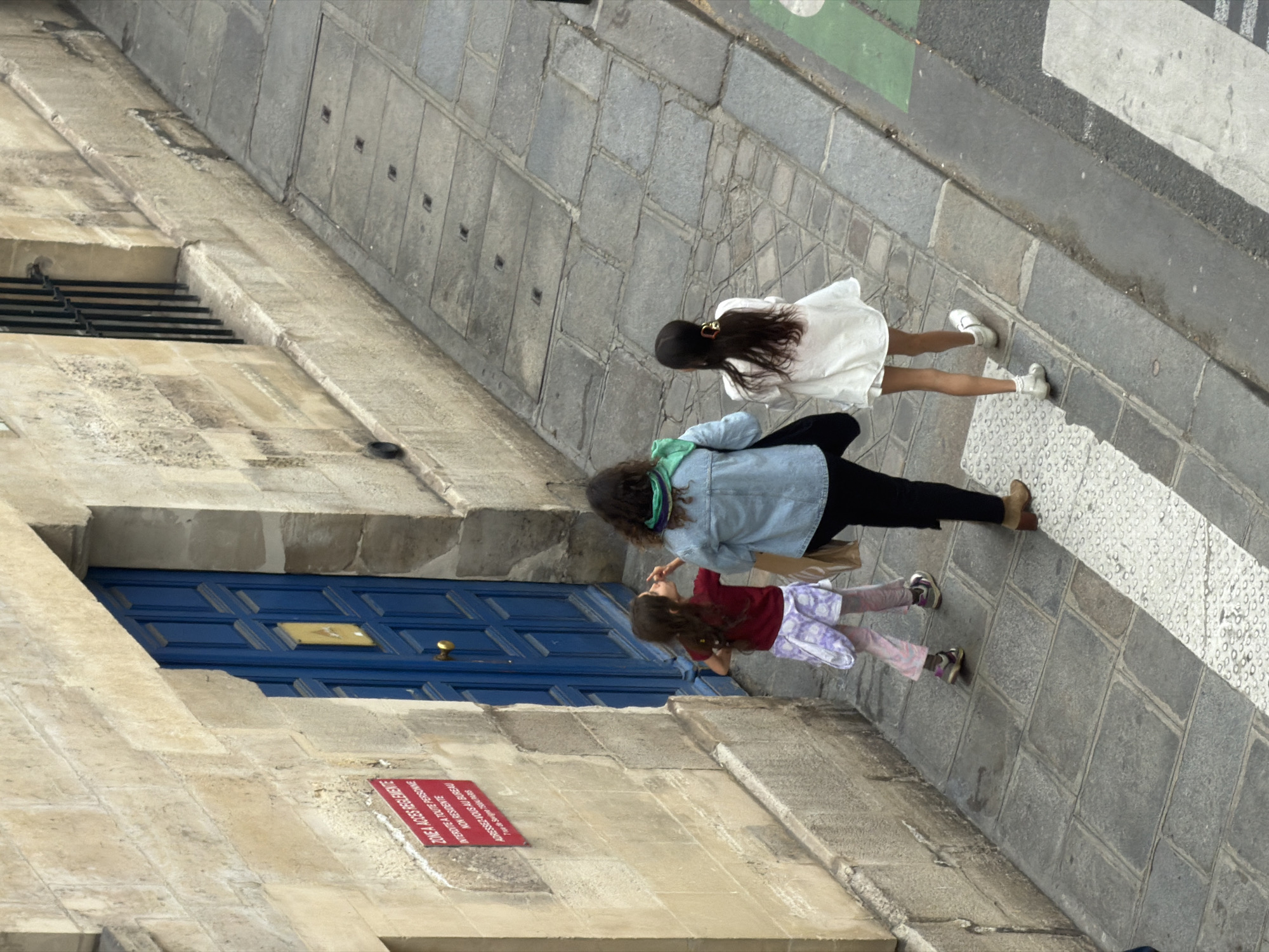 The image shows a street scene in Paris, France, taken from an elevated perspective. Three females of varying ages are walking across a pedestrian crosswalk. The youngest female is wearing a red shirt and patterned pants, while the second is wearing a denim jacket and black pants, and the eldest is wearing a white dress. They are walking in a single direction. To the left of the pedestrians is a building made of stone blocks, with a blue door. A sign is attached to the building near the blue door. The pavement consists of grey stone blocks. A portion of a street is visible above the pedestrians.