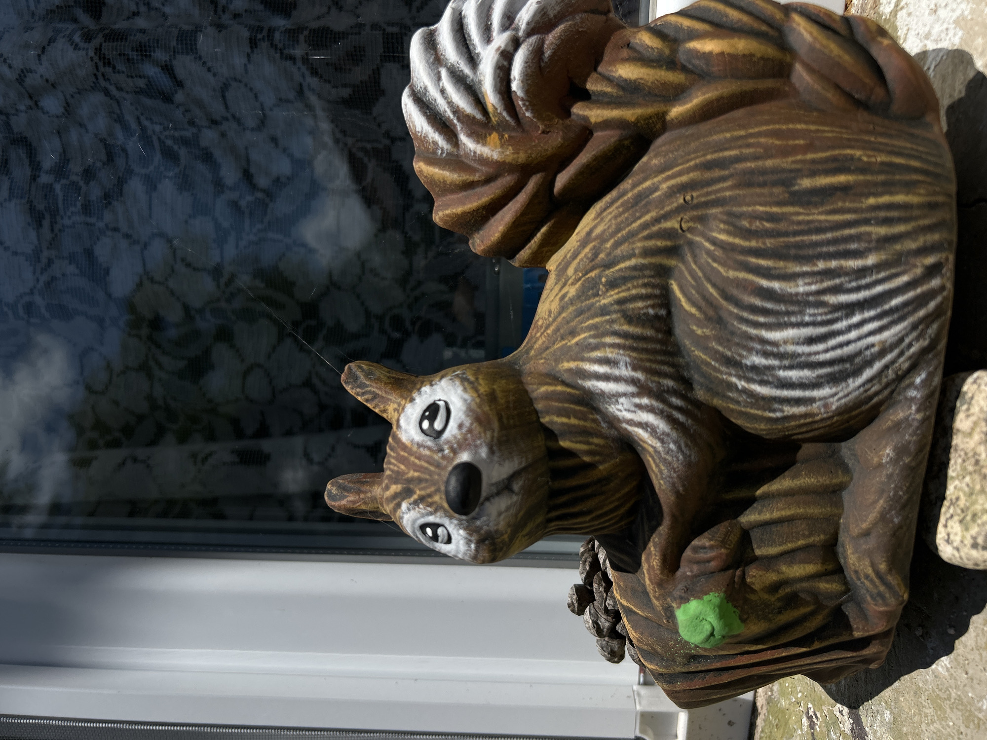 A close-up, low-angle shot features a ceramic squirrel garden ornament positioned outdoors. The ornament is brown with white and yellow accents, designed to mimic a squirrel's fur. It is seated, holding a small green object in its left paw and resting on what appears to be a log or tree stump. Behind the ornament, a window with a lace curtain is visible, reflecting the environment. The foreground shows a stone surface with a few small rocks, suggesting an outdoor garden or patio setting. The overall lighting indicates it is daytime. There are no people or animals present in the image. The location is not specified as Belo pole, Bulgaria.