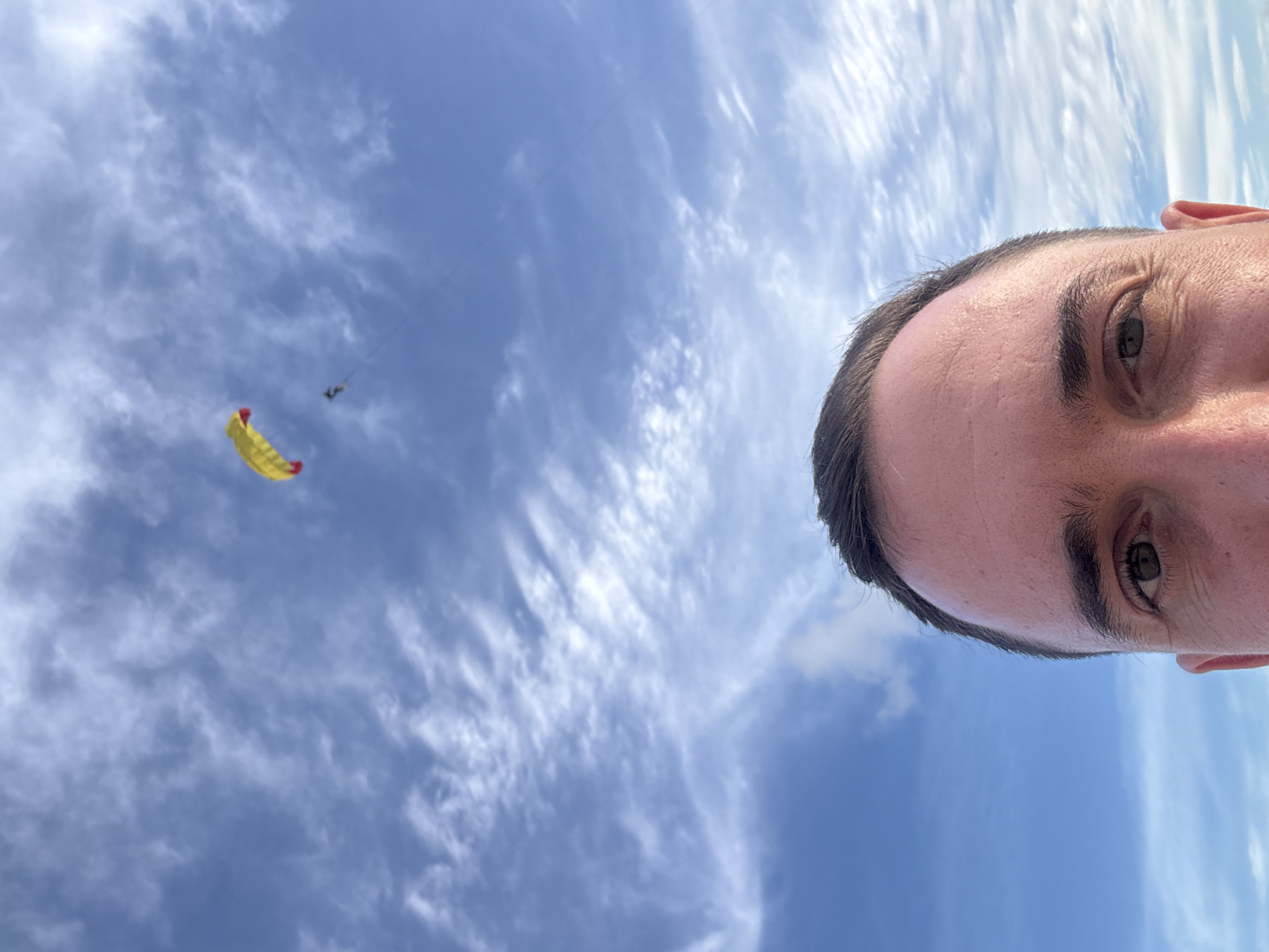 A close-up selfie is taken of a man's face, with his forehead and eyes filling the bottom half of the frame. He has short, dark hair and light skin. His eyes are looking directly at the camera. Above his head, the sky is a vibrant blue with scattered white clouds. A yellow and red kite is visible in the upper right quadrant of the image, with a smaller black figure, possibly a person, suspended below it. The kite and figure are flying high in the sky, suggesting an outdoor activity. The scene appears to be a bright, sunny day, likely in an open area where kite flying is possible. The location is identified as Karon, Thailand, which is known for its beaches and opportunities for outdoor recreation.