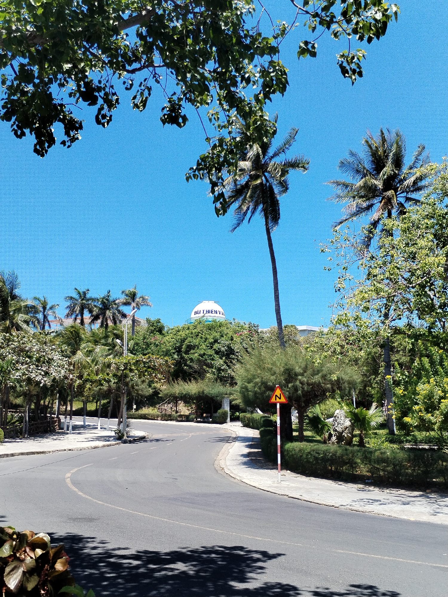 The image shows a street in Bac Nha Trang Ward, Vietnam. The street curves to the left and is surrounded by trees and vegetation. Palm trees are visible, and a white domed building with lettering is seen in the background, atop a hill. A yellow and red triangular road sign with the letter "N" is visible on the right side of the road. The sky is clear and blue.
