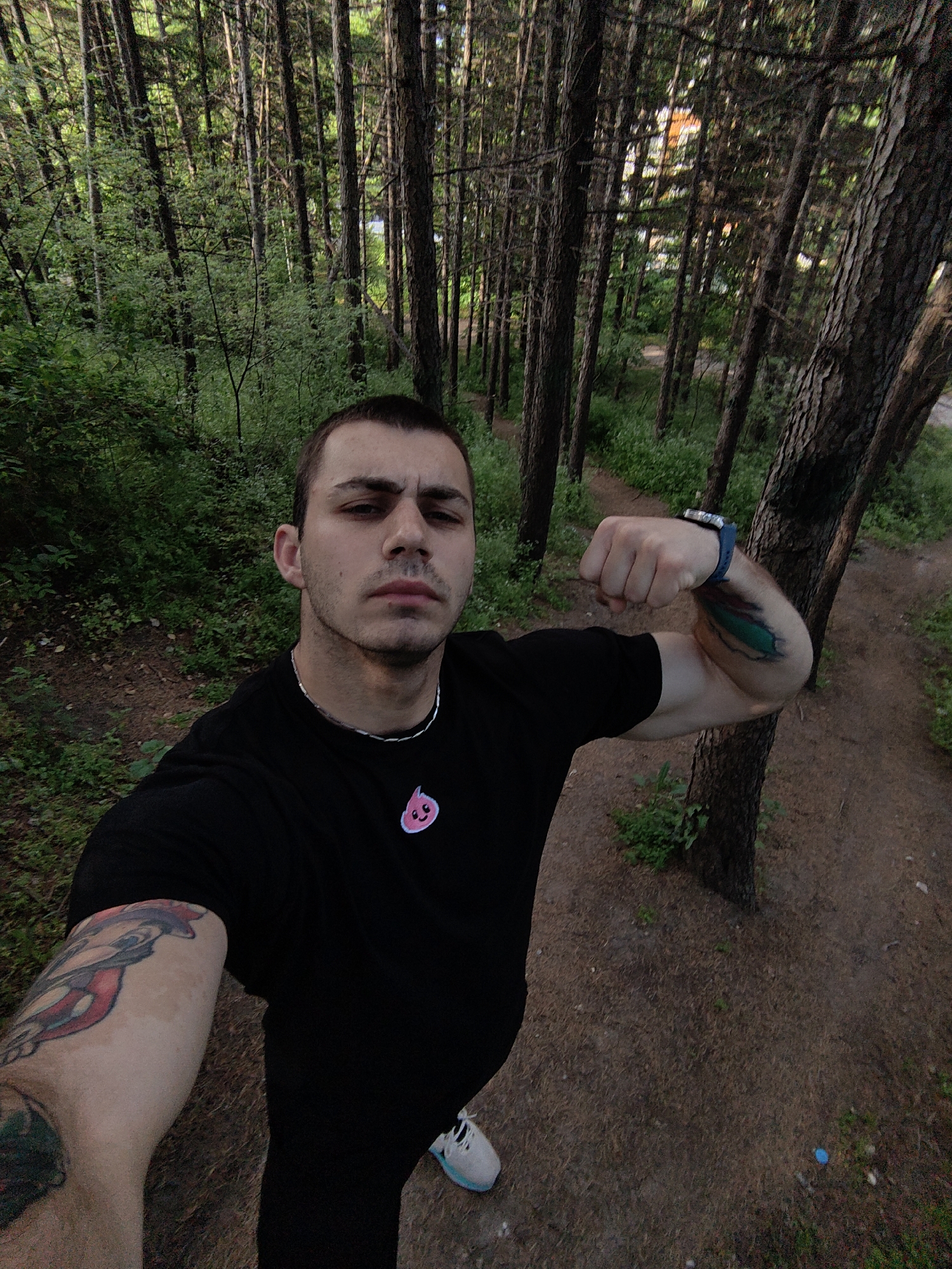 A man is posing for a selfie in a forest in Varna, Bulgaria. He is wearing a black t-shirt with a pink logo, a silver necklace, and a blue watch. He has tattoos on both arms. He is flexing his right bicep. The forest has tall, thin trees and green vegetation. A path is visible behind him.