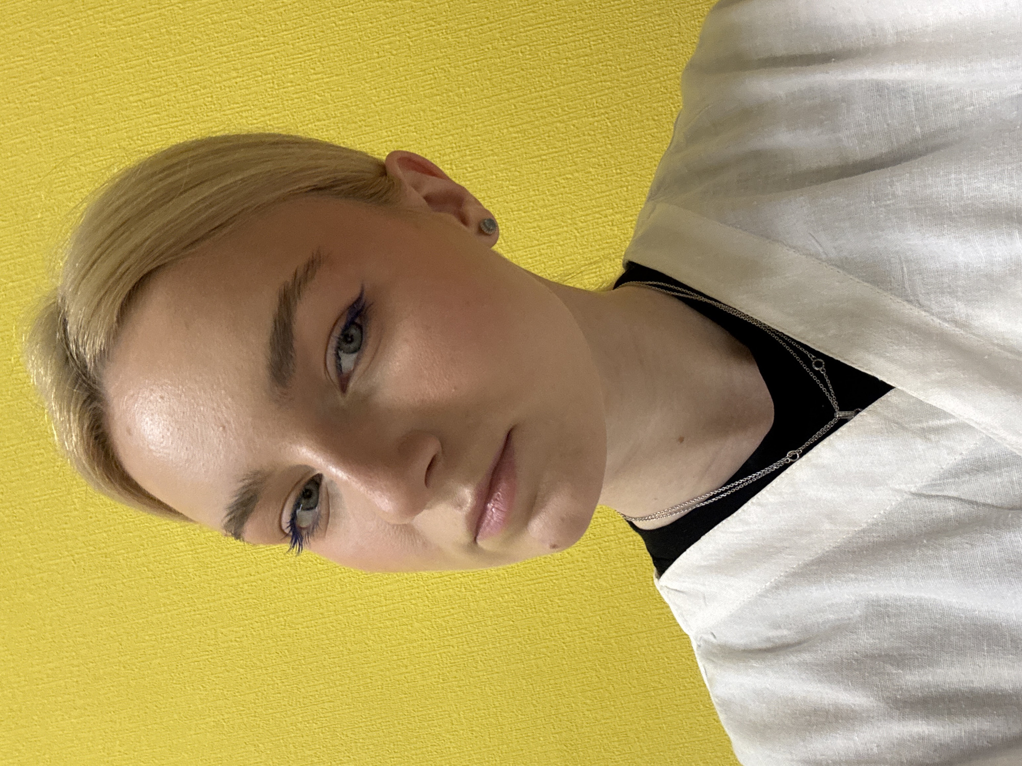 A young woman with blonde hair pulled back is positioned in front of a textured yellow background. She is wearing a white collared shirt over a black garment and silver necklaces. Her makeup includes blue eyeliner and blush. The image is a close-up, focusing on her face and upper body. The location is not specified in the image.