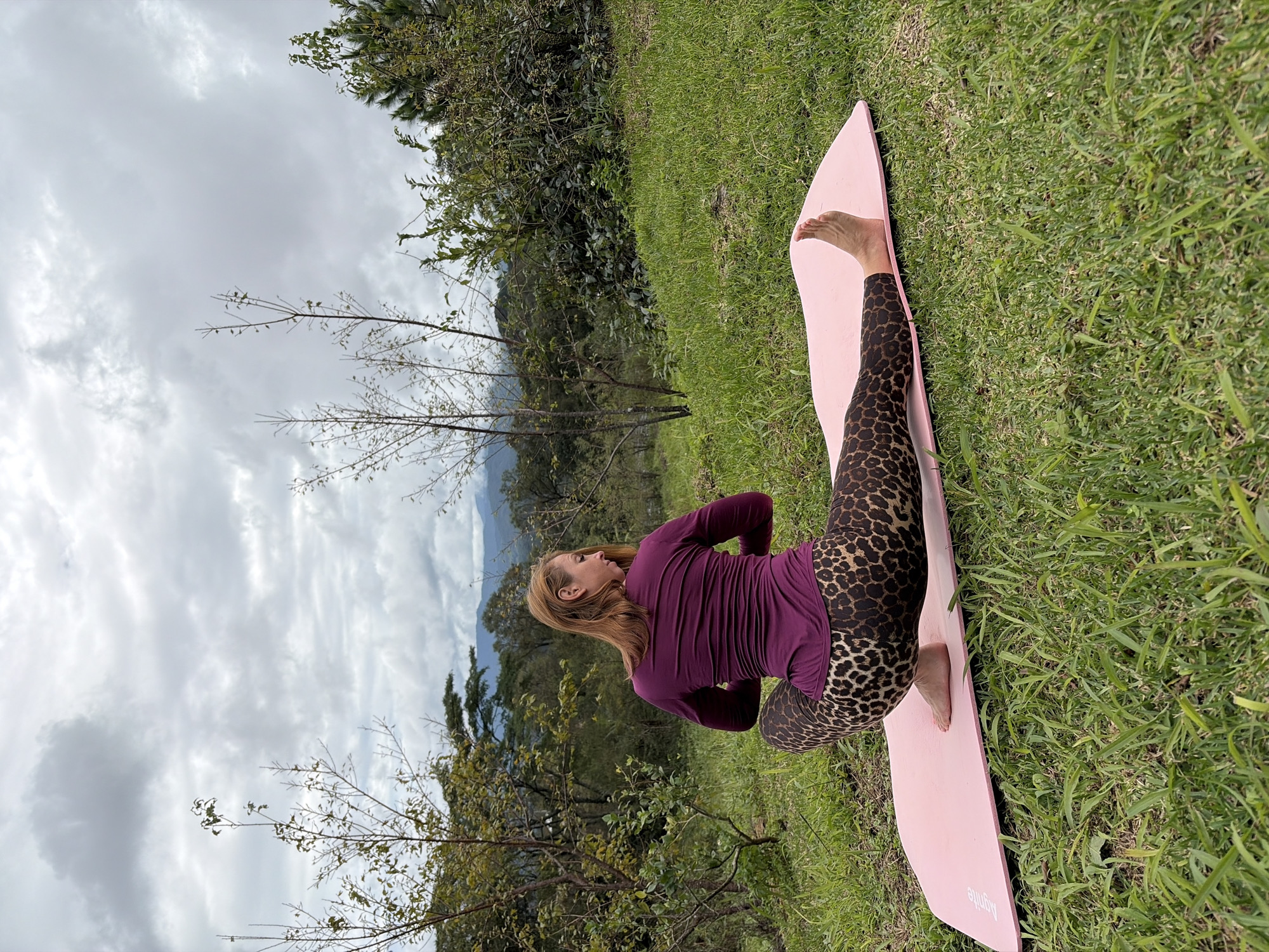 In Ban Khun Wang, Thailand, a woman with auburn hair performs a yoga pose on a pink mat placed on a grassy slope. She is wearing a long-sleeved purple top and leopard-print leggings. Her body is twisted to the left, with her right leg extended forward and her left leg bent with the foot tucked near her hip. Her hands are clasped in front of her chest. The background features lush greenery, scattered trees, and a cloudy sky. In the distance, mountains are visible. The scene is set outdoors, with natural light illuminating the woman and her surroundings.