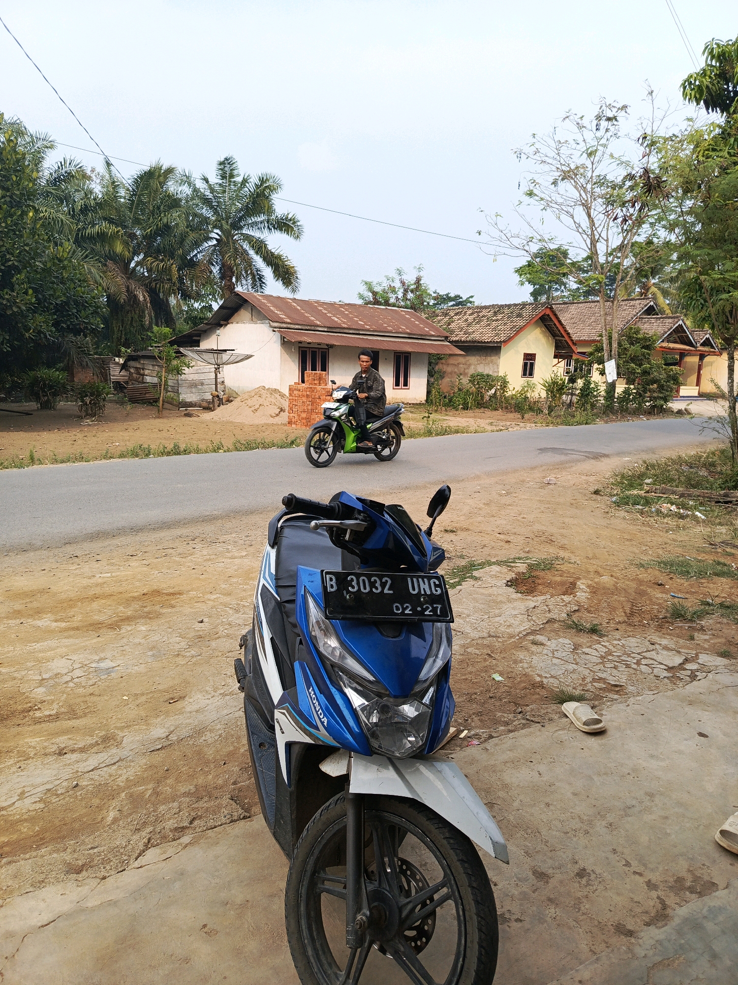 The image depicts a street scene in Megang Sakti I, Indonesia. In the foreground, a blue and white scooter is parked on a dirt and concrete surface. The scooter has a license plate that reads "B 3032 UNG 02-27". Behind the parked scooter, a paved road runs horizontally across the frame. A young man wearing a dark jacket is riding a green and black motorcycle on the road, heading away from the viewer. He is looking forward. To the left of the road, there are several houses with light-colored walls and tiled roofs. Tall palm trees and other vegetation are visible around the houses. The background shows a clear sky with some clouds. The environment appears to be rural or semi-urban, with evidence of construction materials like bricks and sand near one of the houses.