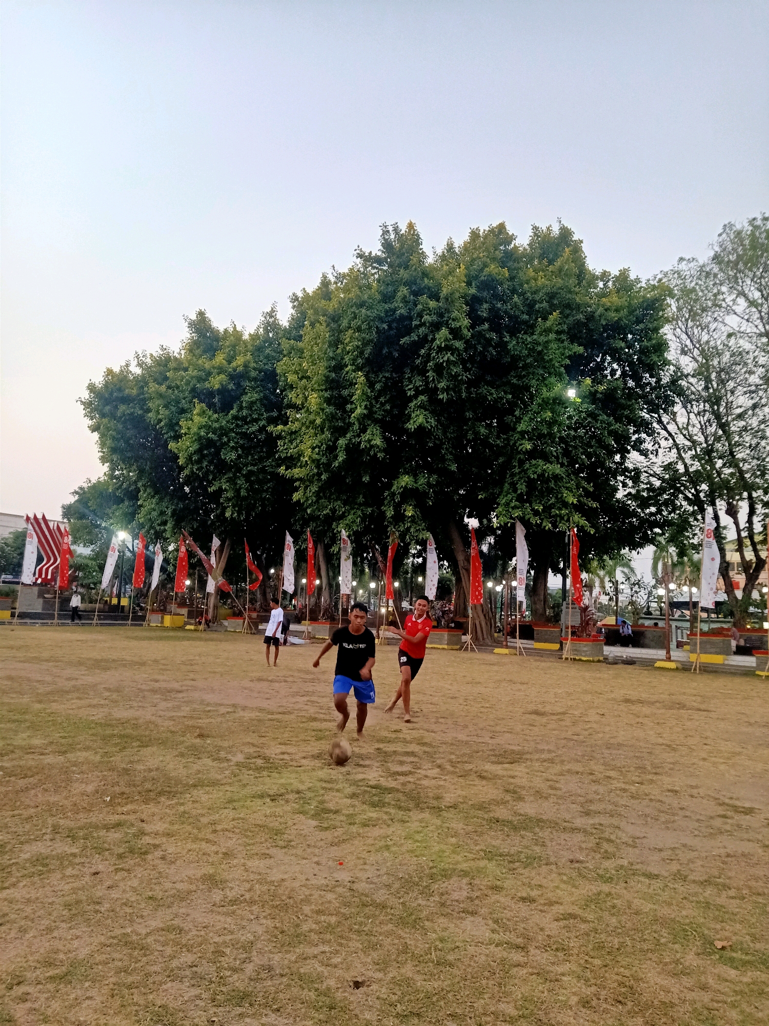 This is an outdoor, eye-level, full-shot photograph of a park in Klaten, Indonesia, where a group of young men are playing football. The sky is a pale, hazy blue, indicating late afternoon or early morning. The ground is covered in dry, yellowish-brown grass, with patches of greener grass interspersed. Large, mature trees with dense green foliage dominate the background, their branches reaching towards the sky.
In the foreground, a young man wearing a black t-shirt with white lettering and blue shorts is in motion, appearing to be kicking or passing a soccer ball. Another young man, wearing a red t-shirt with the number '25' on his left sleeve and black shorts, is also in motion, facing the first man and seemingly engaged in the game. A third young man, in a white t-shirt and dark shorts, is visible further in the background, also appearing to be participating. Scattered around the park are numerous flags, mostly red and white, attached to poles. These flags are arranged in rows along the perimeter of the playing area and further back near the trees. Some structures, possibly benches or decorative elements, are visible behind the flags. The overall impression is a casual sports activity taking place in a well-maintained park setting within the city of Klaten.