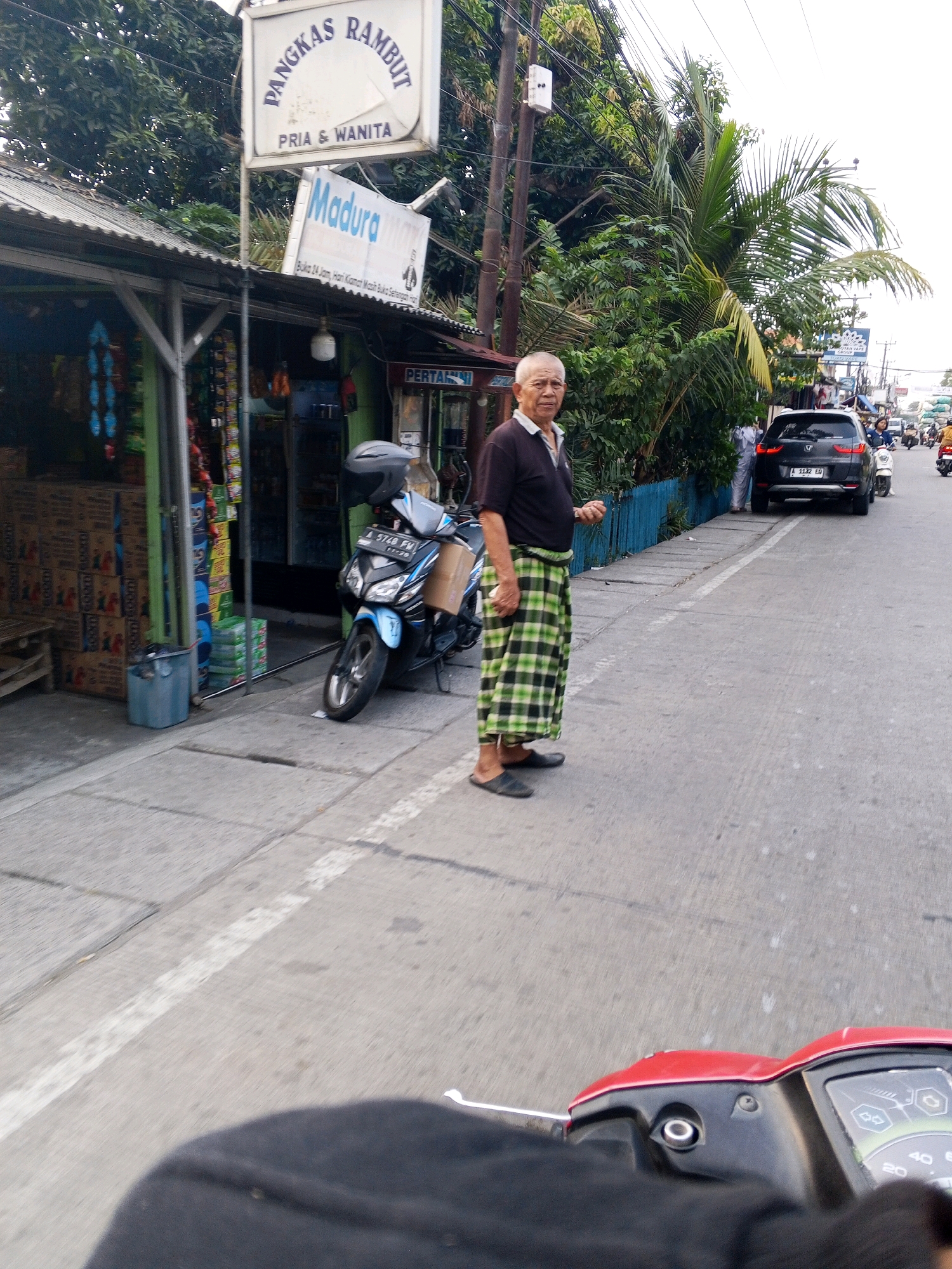 The image captures a street scene in Ciruas, Indonesia. In the foreground, a person is partially visible, their hand and a red motorcycle dashboard are the most prominent features. The motorcycle is stopped. To the left, a small shop with a corrugated metal roof displays various goods, including stacked boxes and hanging items. A blue trash bin is situated on the sidewalk in front of the shop. A blue and black scooter is parked near the shop.
In the middle ground, an elderly man stands on the sidewalk wearing a dark polo shirt and a green and black checkered sarong. He is facing towards the viewer, with his left hand slightly raised. Behind him, a black SUV is parked on the street, with its license plate clearly visible. The street is paved and has a white line marking the lane. Several other vehicles, including scooters and a white car, are visible further down the street, indicating ongoing traffic.
In the background, lush green trees and vegetation line the street. A white sign with Indonesian text "PANGKAS RAMBUT PRIA & WANITA" (Hair Salon for Men & Women) and another smaller sign below it that reads "Madura" are prominently displayed above the shop. Utility poles with wires run along the side of the road. The sky is overcast.