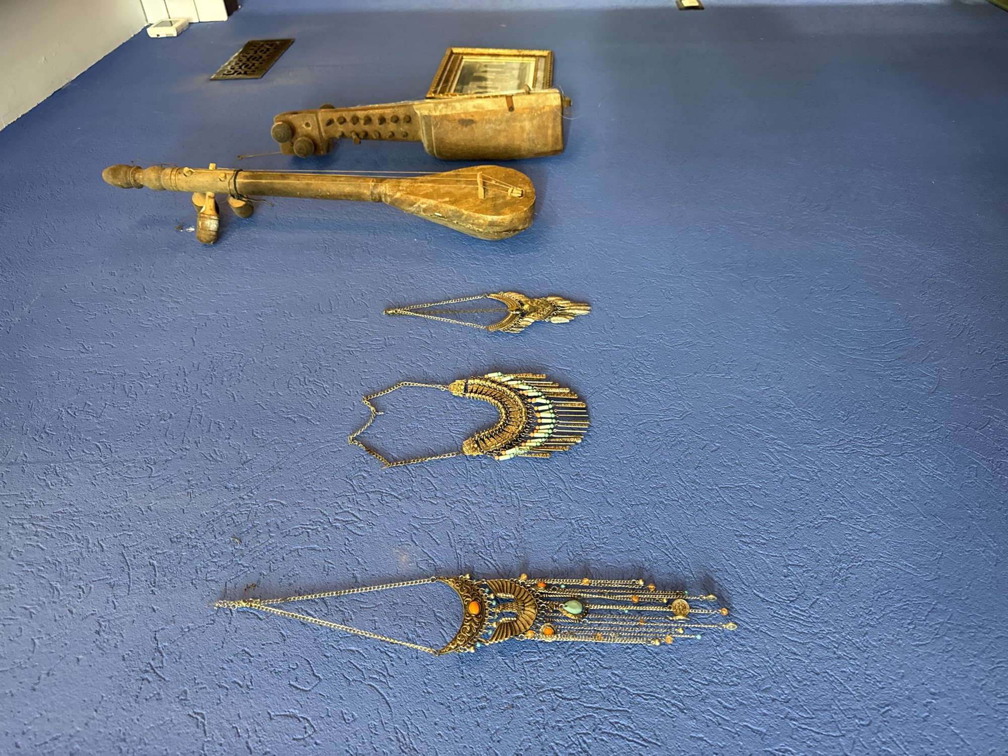 The image shows a blue textured wall in what appears to be a shop or home in Hof HaCarmel Regional Council, Israel. The wall is adorned with several decorative items. On the left, there are three long, ornate necklaces, each with a chain and a pendant. The leftmost necklace is the longest and features a large, golden eagle-like emblem with dangling chains and beads in orange and turquoise. The middle necklace is shorter and has a semicircular pendant with multiple layers of gold fringe and small green and orange beads. The smallest necklace on the far right has a golden, feather-like pendant. To the right of the necklaces, two wooden stringed instruments, possibly lutes or similar folk instruments, are hung vertically. The instrument on the left is lighter in color and appears older. The instrument on the right is darker and has a more pronounced body. Between these instruments and further to the right, a small, gilded framed picture is hung on the wall. The overall impression is one of a decorative display of cultural or artistic items.