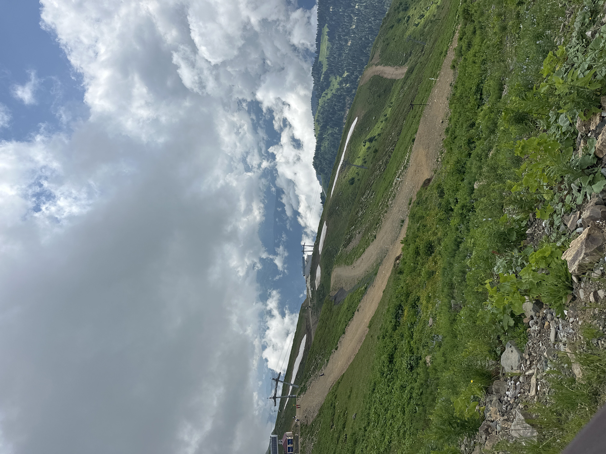 An aerial view captures a mountainous landscape with a cloudy sky. In the foreground, a rocky, green slope is covered with sparse vegetation. Ascending the mountain, a ski resort path is visible, winding its way upwards. Patches of snow dot the grassy slopes, interspersed with ski lift pylons. Further up, ski lifts and structures are present. The background reveals rolling hills covered in dense green forests, with more clouds obscuring the distant peaks. The scene depicts a summer day in a mountainous ski resort.