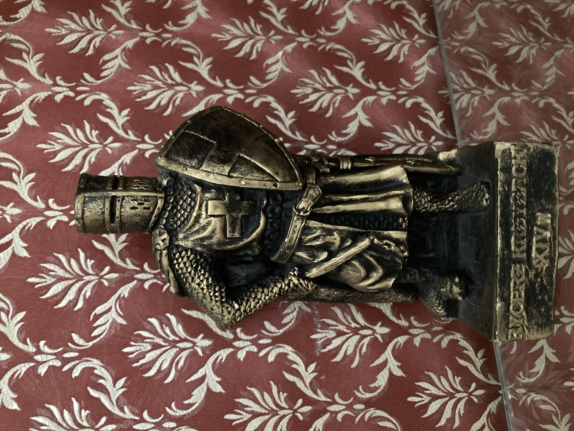 The image shows a close-up, eye-level, full-shot of a golden statue of a knight in armor, standing against a red and white damask wallpaper. The knight is depicted in a stoic pose, with his left hand on his hip and his right hand holding a shield. He wears a helmet, chainmail, and a surcoat with a cross emblem. A sword is visible at his side, and a dagger is tucked into his belt. The base of the statue has an inscription that reads "RYCERZ KRZYSZTOF XIV." The statue is placed on a reflective surface, possibly glass, which partially shows the wallpaper pattern below. The lighting appears to be indoor, casting subtle shadows on the statue and the wallpaper.