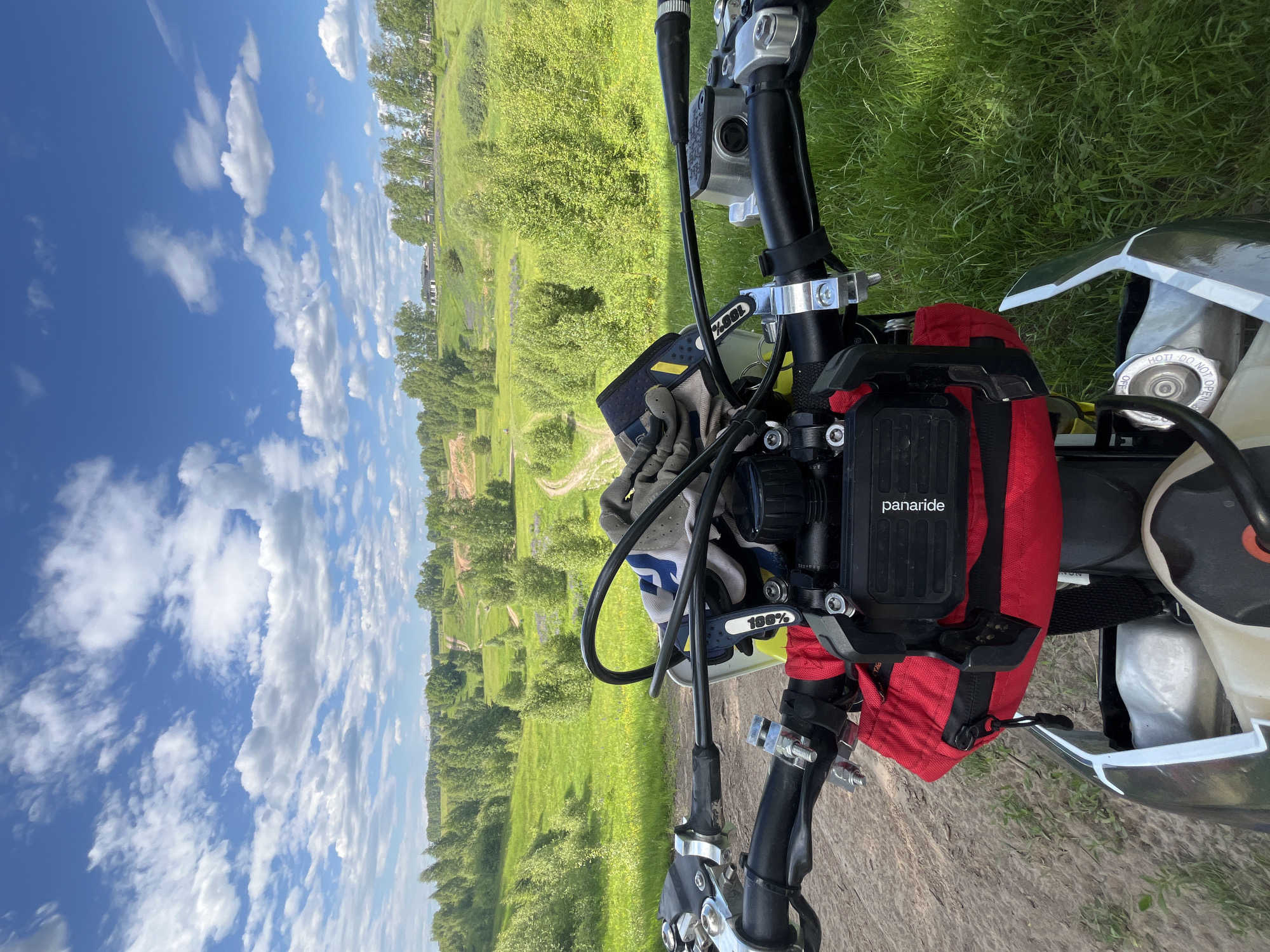 The image depicts a first-person perspective from atop a motorcycle, stationary on a dirt path in Новочувашский, Russia. The motorcycle's black handlebars, silver clamps, and control levers are prominent in the lower foreground. A pair of grey and blue gloves, marked with "100%", rests on the handlebars. Below the handlebars, a red fabric bag is attached, and a black electronic device or case, labeled "panaride", is mounted above it. A portion of the motorcycle's light-colored fuel tank is visible, featuring a fuel cap with the inscription "DO NOT OPEN HOT".
The scene unfolds into a rural landscape characterized by rolling green hills. A winding dirt track extends into the midground, flanked by lush green grass and numerous deciduous trees with dense foliage. Patches of purple flora are visible within the grassy areas on the hillsides. In the distance, several light-colored buildings, presumed to be houses, are situated on a ridge, partially obscured by trees. To the left, a distinct area of disturbed ground or a quarry with lighter soil is visible on a hillside. The sky above is a clear blue, dotted with scattered white cumulus clouds. The immediate ground beneath the motorcycle is a mix of dirt and sparse green grass, transitioning to thicker grass on the right.
