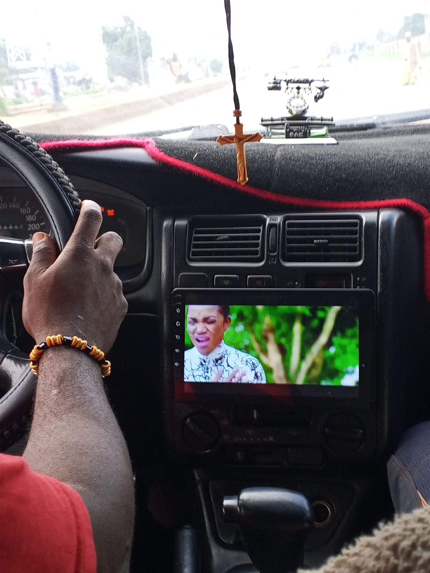 The image shows the interior of a car, likely in Bukuru, Nigeria. A person's hand, wearing a bracelet made of black and gold beads, is visible on the steering wheel. The dashboard features a car stereo displaying a video of a woman, and various car controls. A decorative crucifix hangs from the rearview mirror, accompanied by a small figurine of a rotary telephone. The overall scene suggests a common vehicle interior, possibly encountered in urban or semi-urban environments in Nigeria.