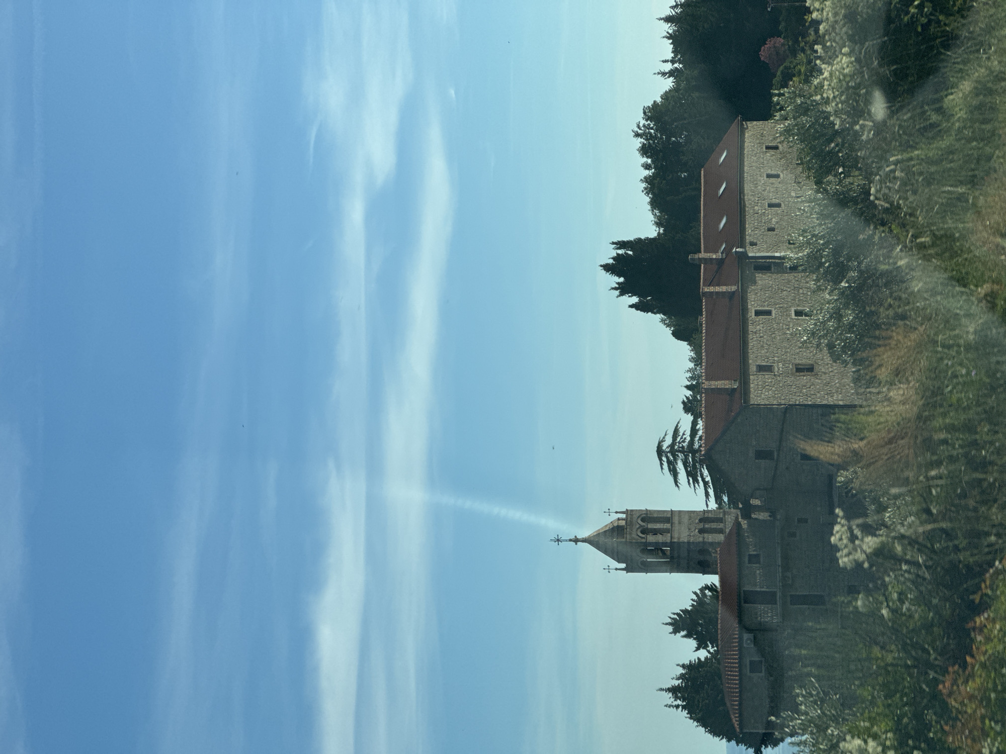 This is a vertical, eye-level, outdoor, medium shot of a stone monastery in Perazića Do, Montenegro, under a clear blue sky with scattered wispy clouds. The monastery features a prominent bell tower with arched openings and a cross at its apex. A streaky cloud formation extends upwards from the tower, creating a column of light. The main monastery building is constructed of stone with red tiled roofs and numerous windows. It is surrounded by dense green trees, including cypress and other evergreens, and lush vegetation in the foreground. The lighting suggests it is daytime.