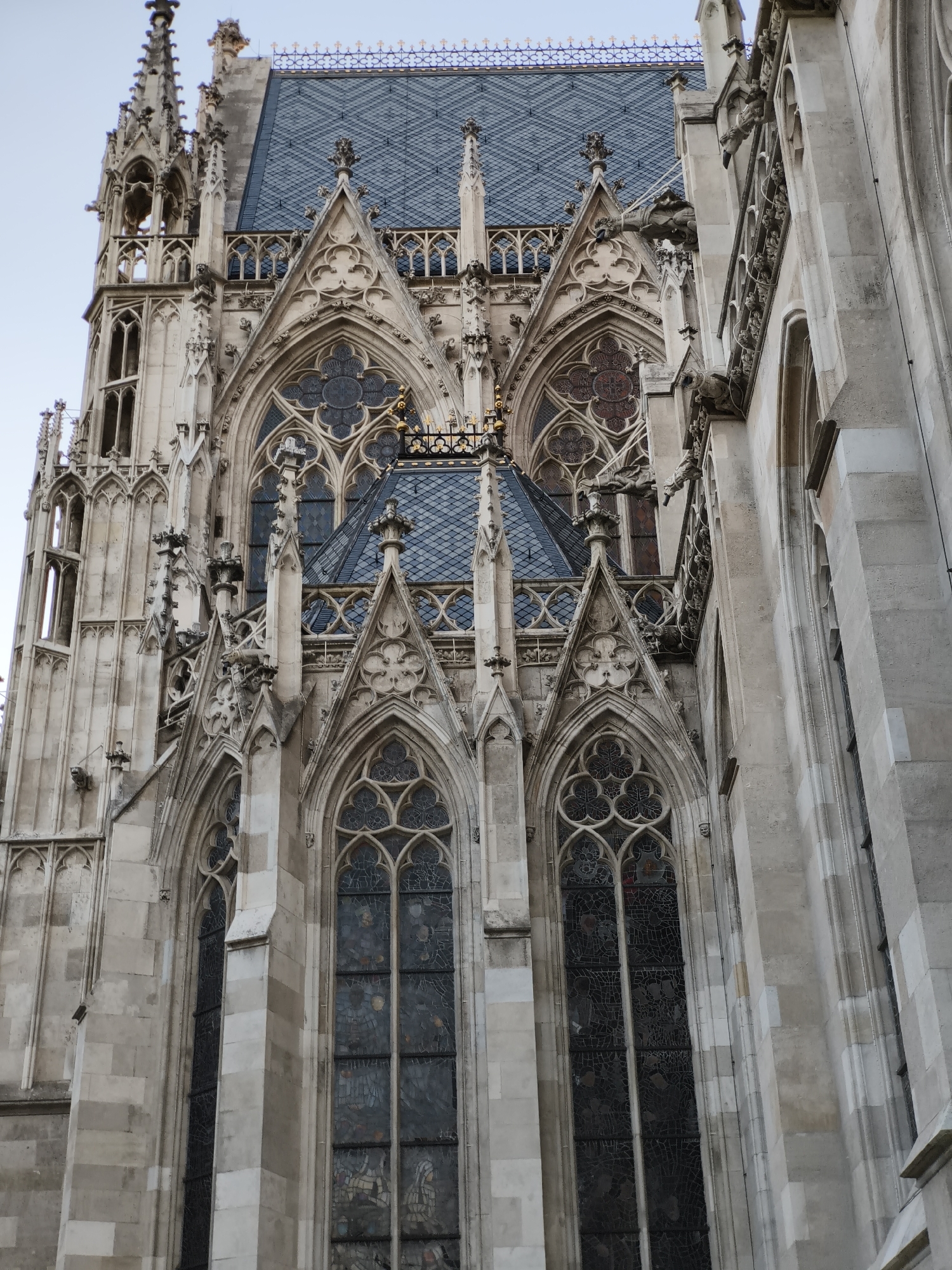 This is a low-angle, medium shot of the exterior of St. Stephen's Cathedral in Vienna, Austria. The architecture is Gothic, with elaborate stonework, pointed arches, and decorative tracery visible on the facade. Several tall, narrow stained-glass windows are prominent, some with intricate geometric and figurative designs. The roof is covered in dark, patterned tiles, and ornate spires and gargoyles are visible on the upper portions of the building. The sky above is a pale, overcast blue. The image focuses on the detailed craftsmanship and imposing scale of the cathedral.