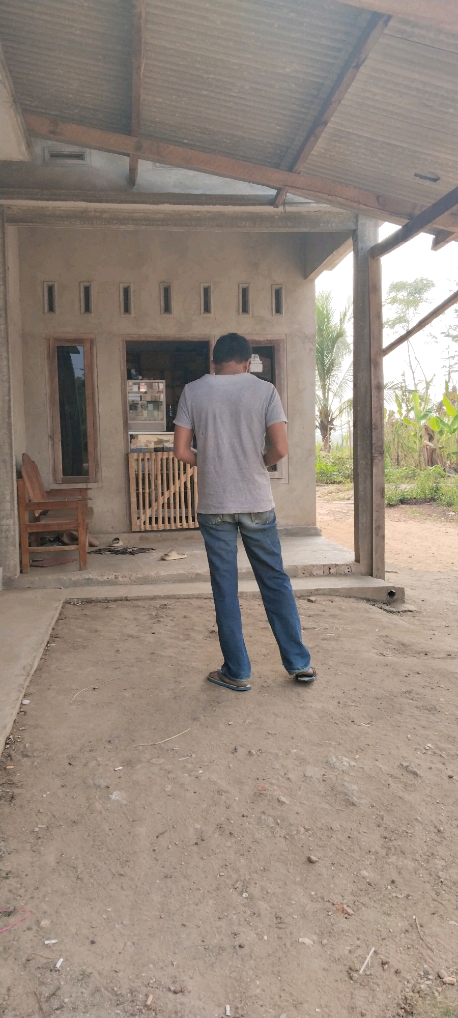 A man stands with his back to the camera, facing a small, open-fronted building. He is wearing a grey t-shirt and blue jeans. The building has a corrugated metal roof supported by wooden beams and a concrete foundation. A wooden chair is visible to the left of the building entrance. The ground is dirt and gravel. The background shows vegetation, including palm trees and what appear to be banana plants. The scene is likely in a rural or semi-rural area of Tanjung Ratu Ilir, Indonesia.