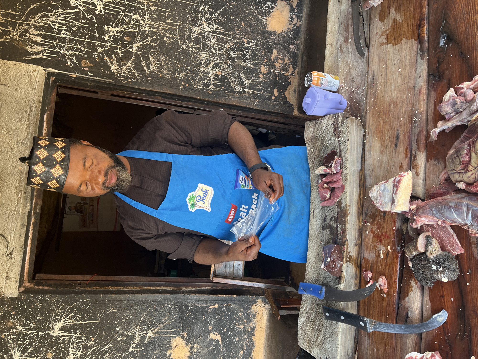A medium shot shows a Black man wearing a dark brown traditional outfit and a blue apron standing in front of a doorway. He wears a black and gold patterned cap. The man is holding a clear plastic bag in his hands, with his fingers inside the bag. He is looking down at the bag. In front of him is a wooden butcher's block with several cuts of raw meat, knives, and a purple pitcher, and a can of Maltina soda. The background shows a dark doorway with a poster on the wall and rough, textured walls with white paint splatters. The scene appears to be an outdoor market stall in Jos, Nigeria, given the presence of the traditional attire, the market setting, and the Maltina can, which is a popular beverage in Nigeria.